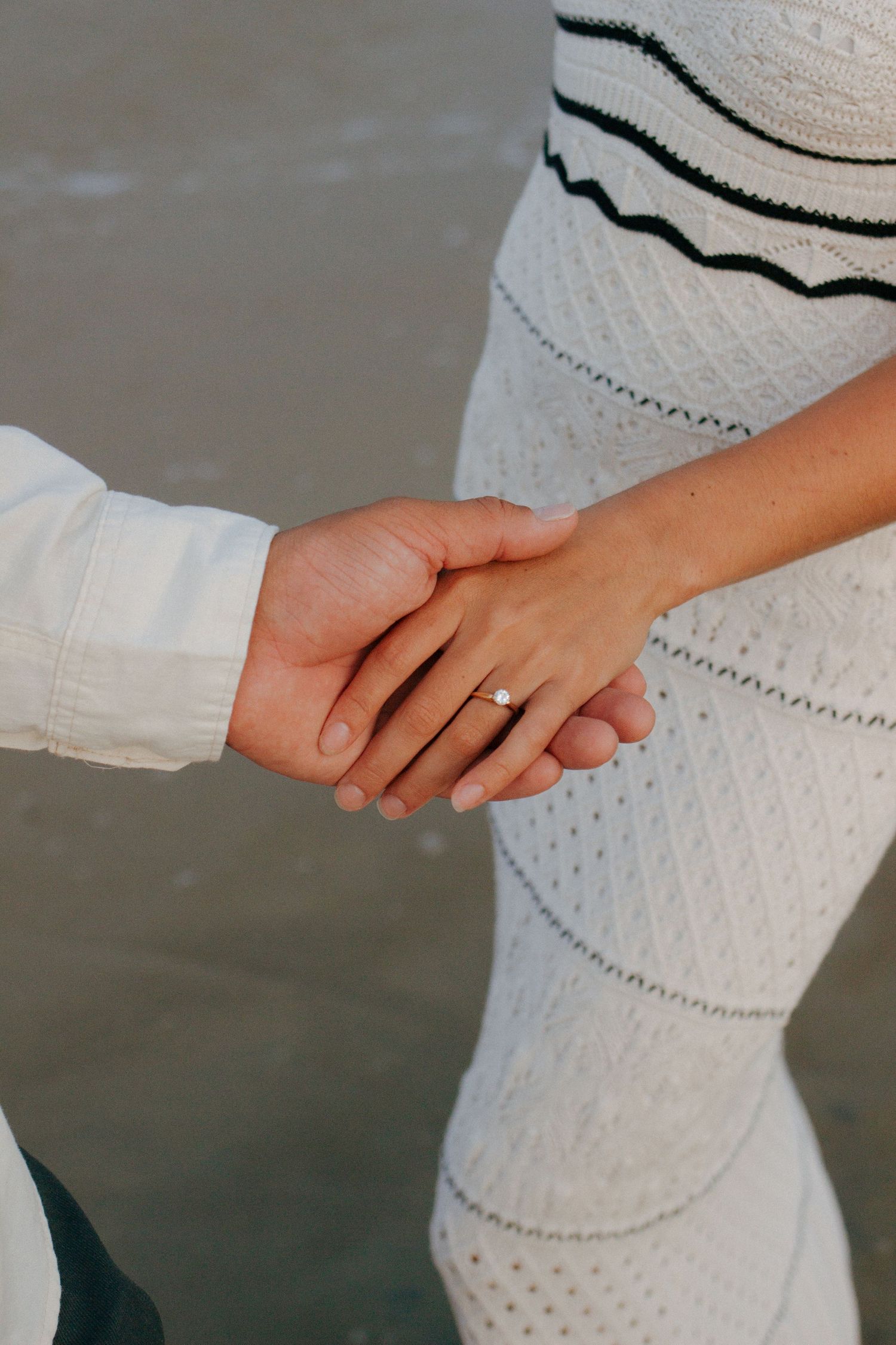Close-up of couple holding hands on beach, woman wearing a diamond engagement ring and white dress.