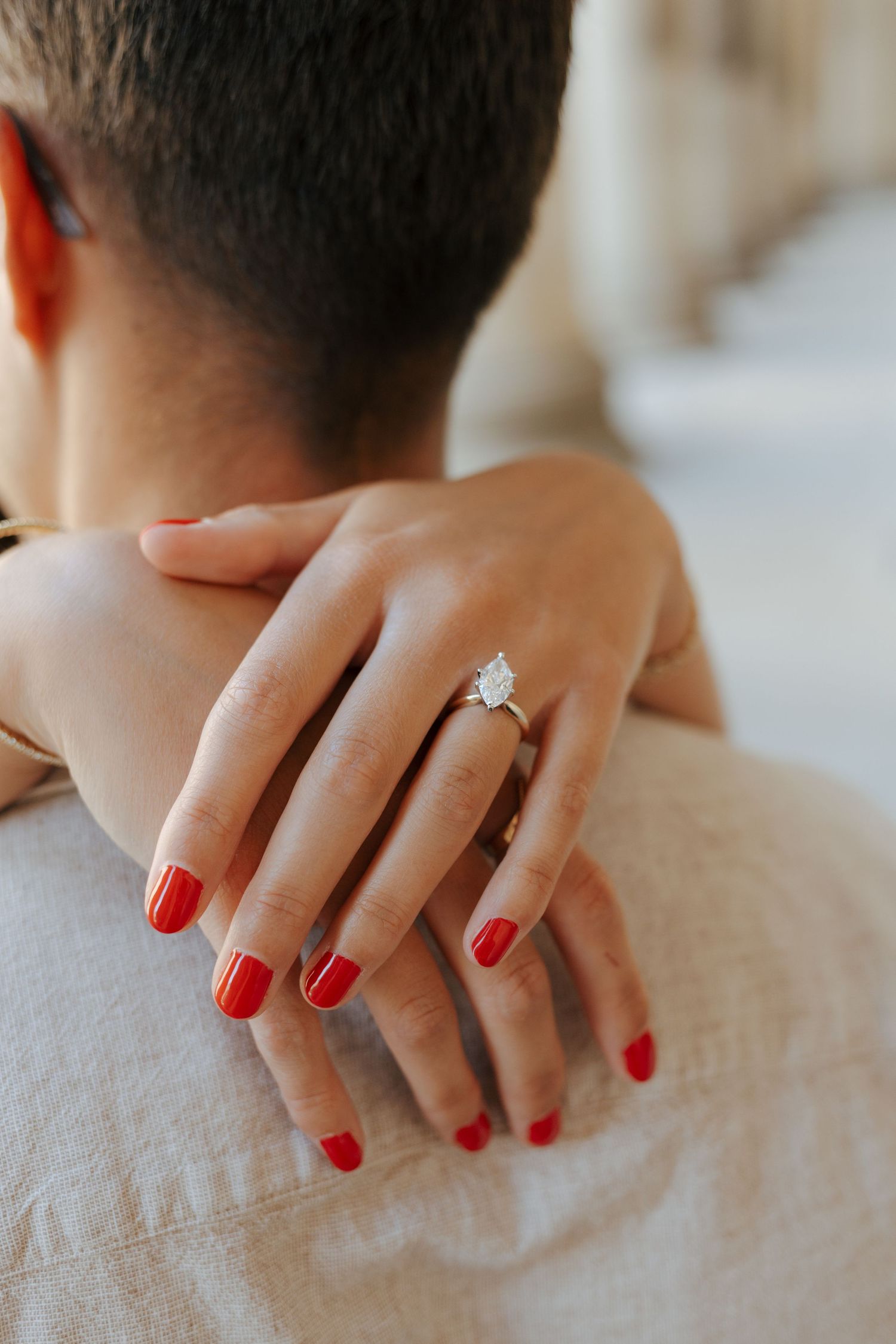 Close-up of a woman's hand with red nails wearing a pear-shaped diamond engagement ring resting on a man's shoulder.