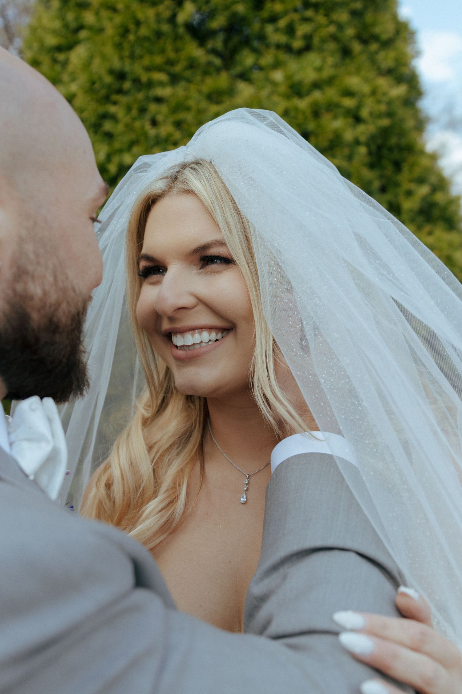 Smiling blonde bride under white veil gazing up at groom outdoors near green hedges.