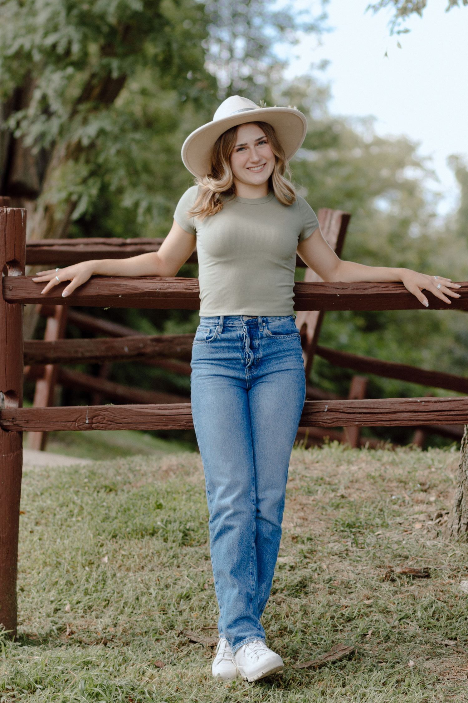 Young woman in sage green top, jeans, and wide-brim hat leaning against a rustic wooden fence outdoors.