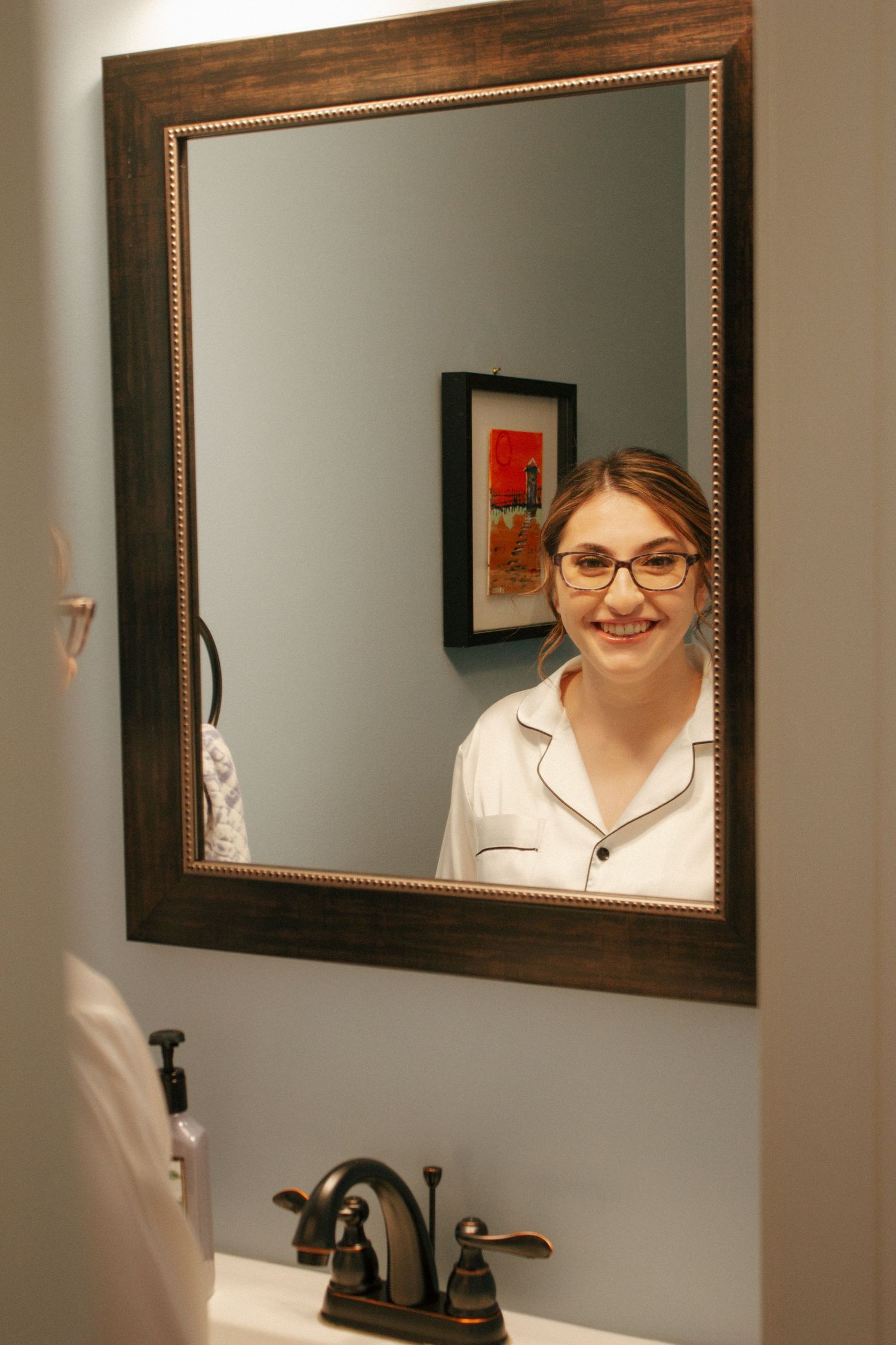 Young woman with glasses smiling at her reflection in a dark wood-framed bathroom mirror.