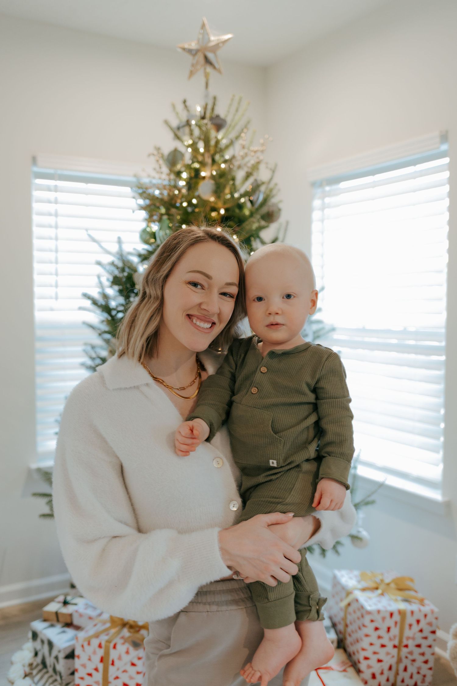 A smiling mother holds her baby boy in front of a lit Christmas tree with wrapped gifts.
