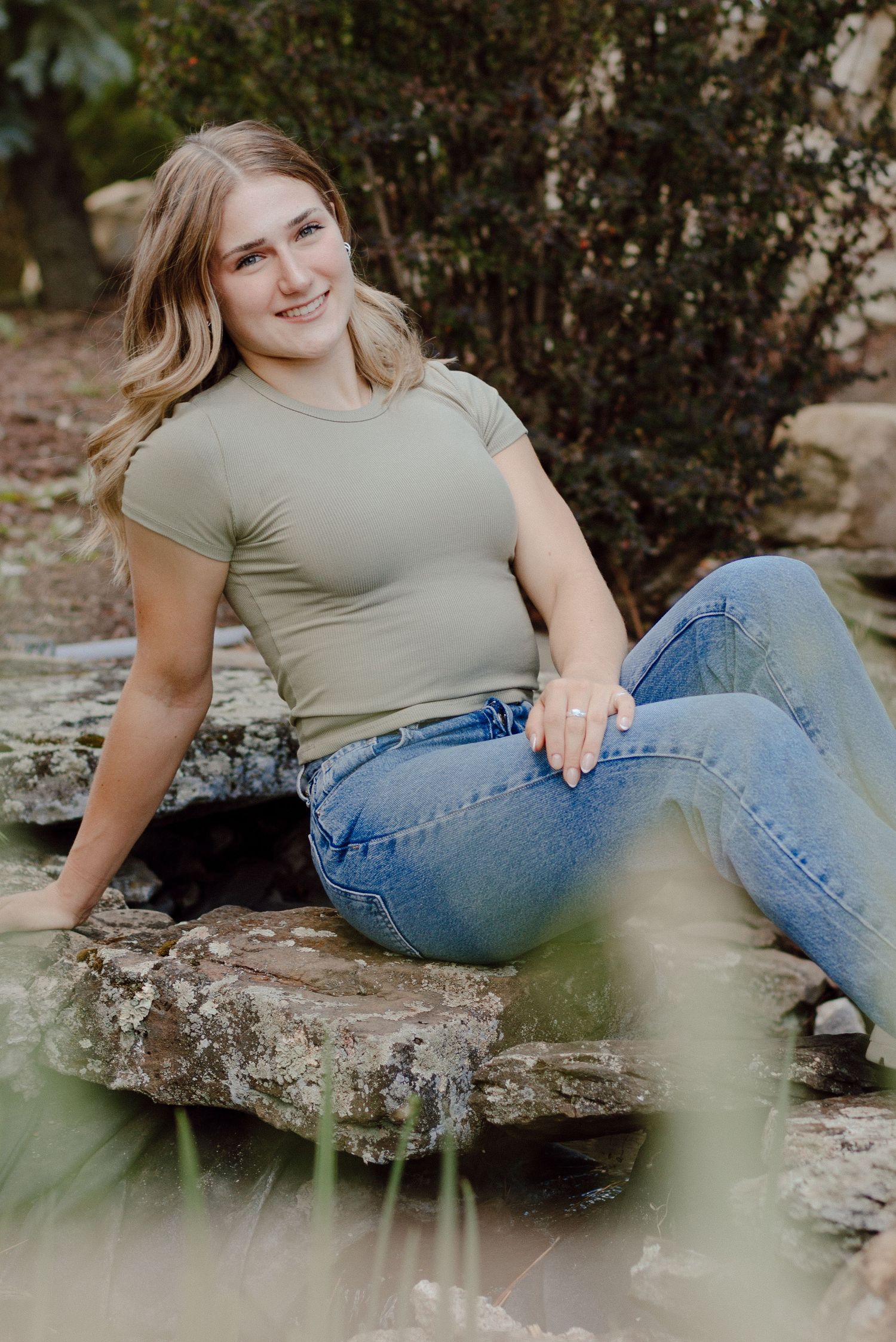 Smiling blonde teen in sage green top and jeans sitting on stone steps outdoors.