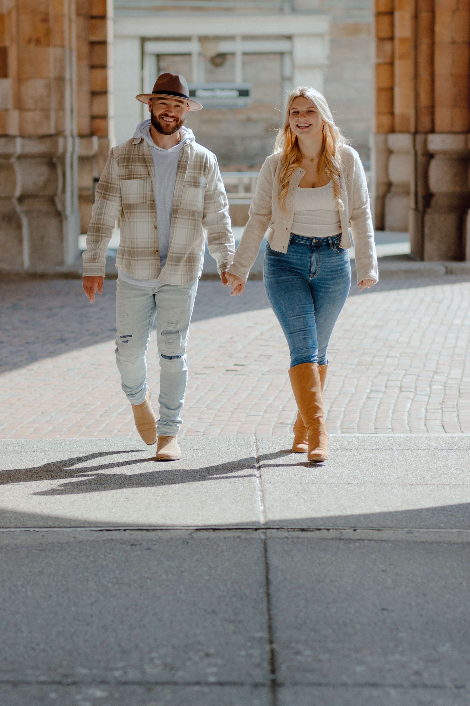 Smiling engaged couple walking hand in hand on a sunny brick plaza, woman wearing brown boots and jeans.