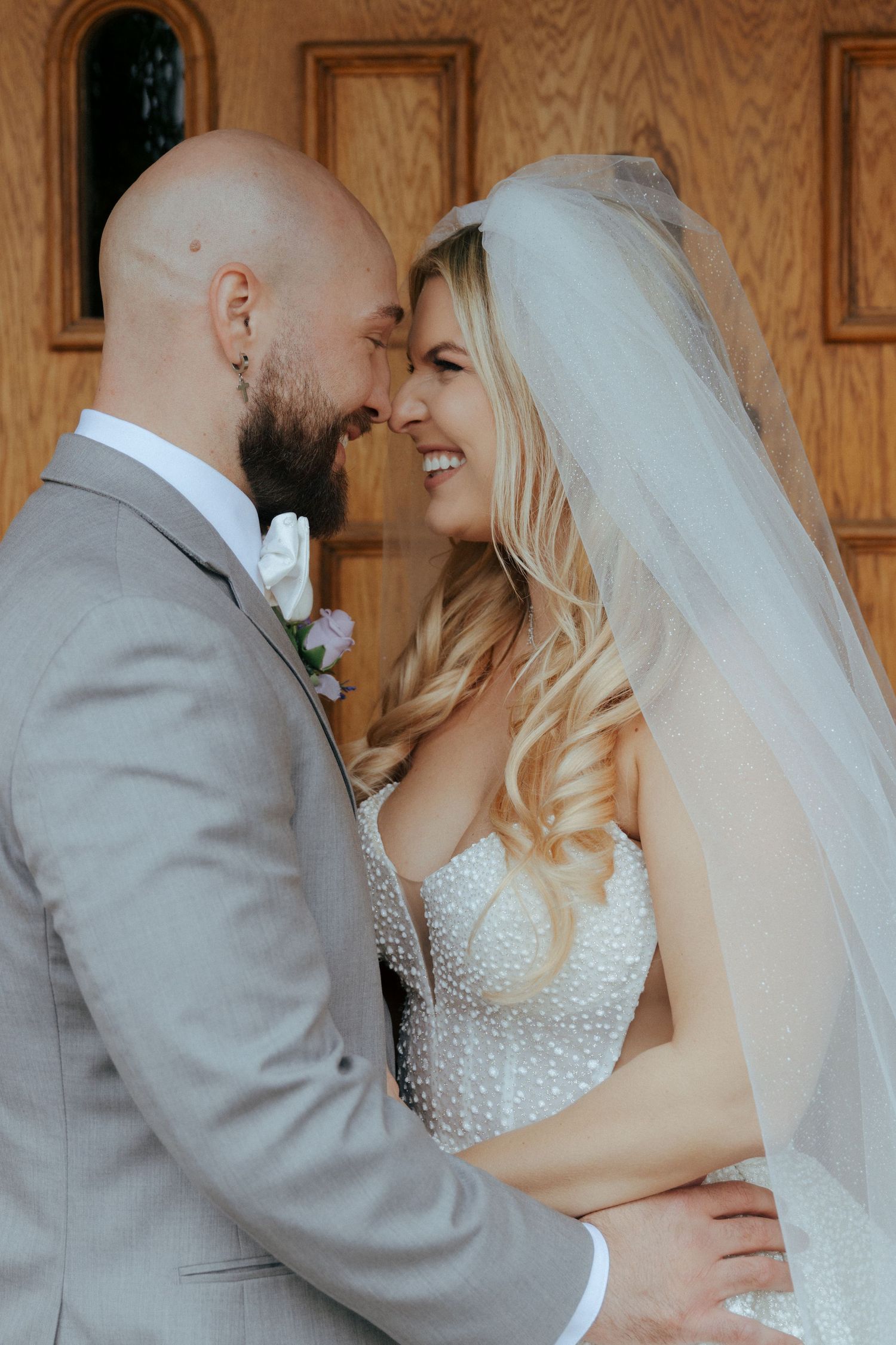 Smiling bride and groom touching foreheads in an intimate moment outside wooden doors on their wedding day.