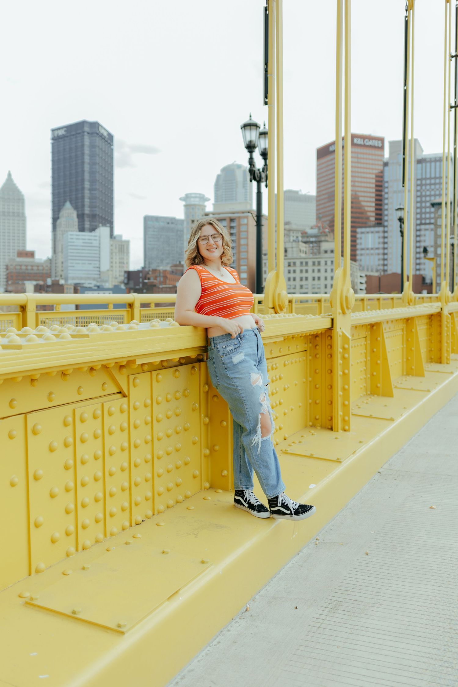 Smiling young woman in orange top and ripped jeans leans on yellow bridge with Pittsburgh skyline.