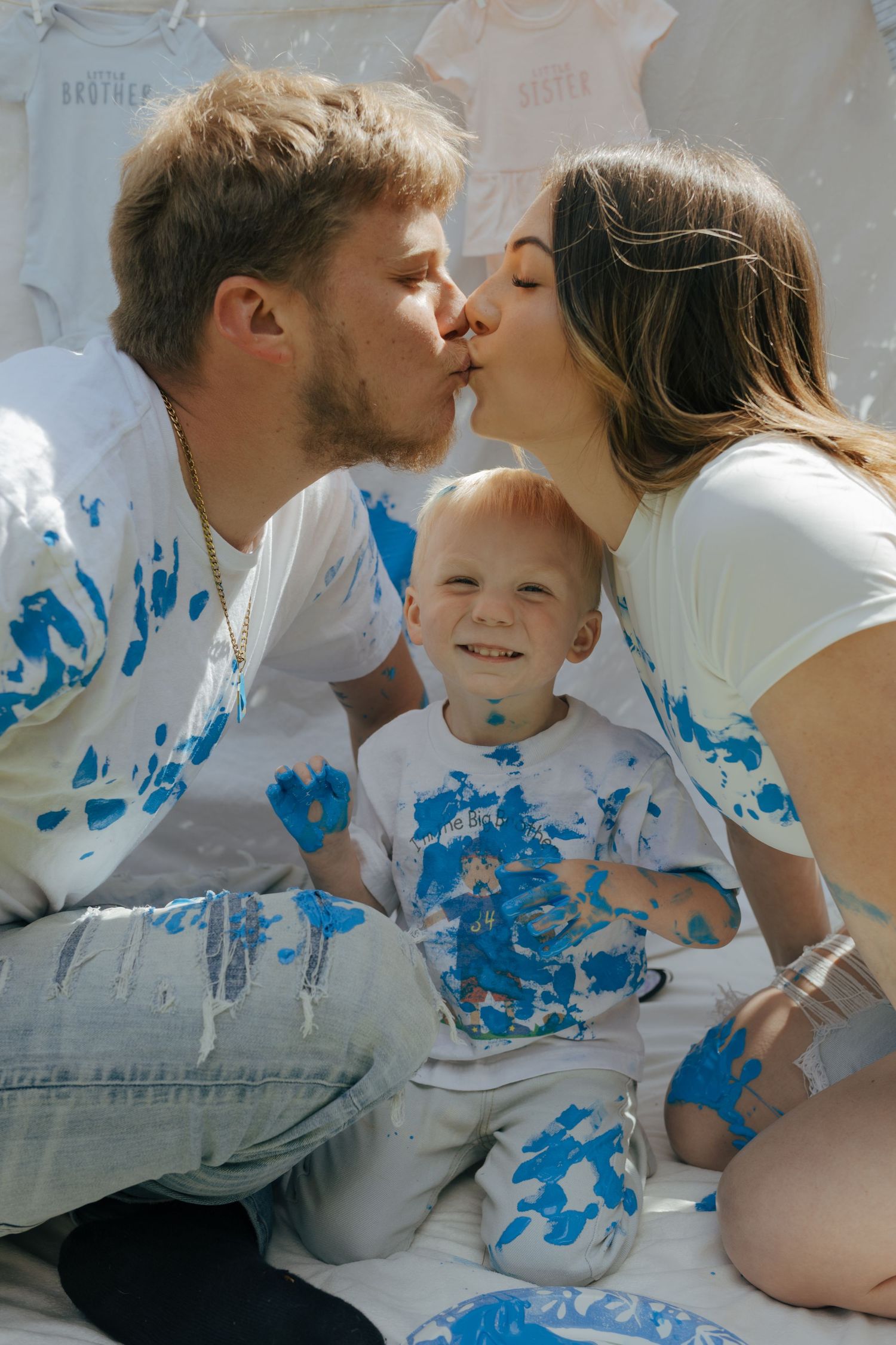 Parents kissing with blue paint on shirts while smiling toddler sits between them.