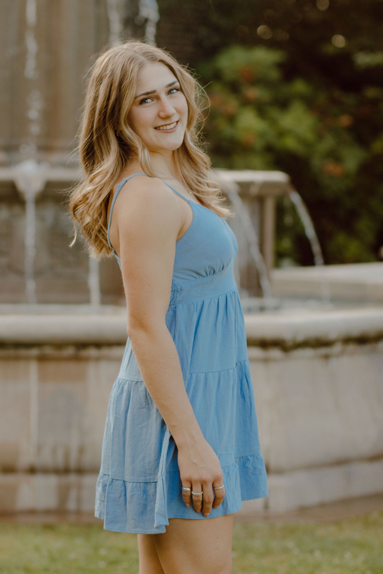 Smiling young woman in a blue sundress posing near a stone fountain in a garden setting.