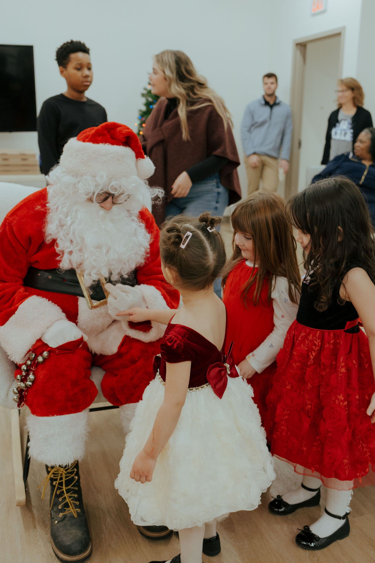 Santa Claus kneels to greet young girls in festive red and white dresses at a Christmas holiday party.