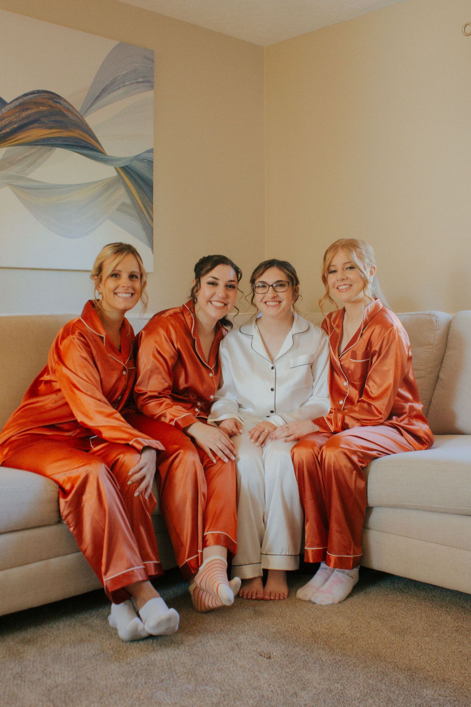 Bride in white satin pajamas poses with three bridesmaids in orange satin pajamas on a couch.