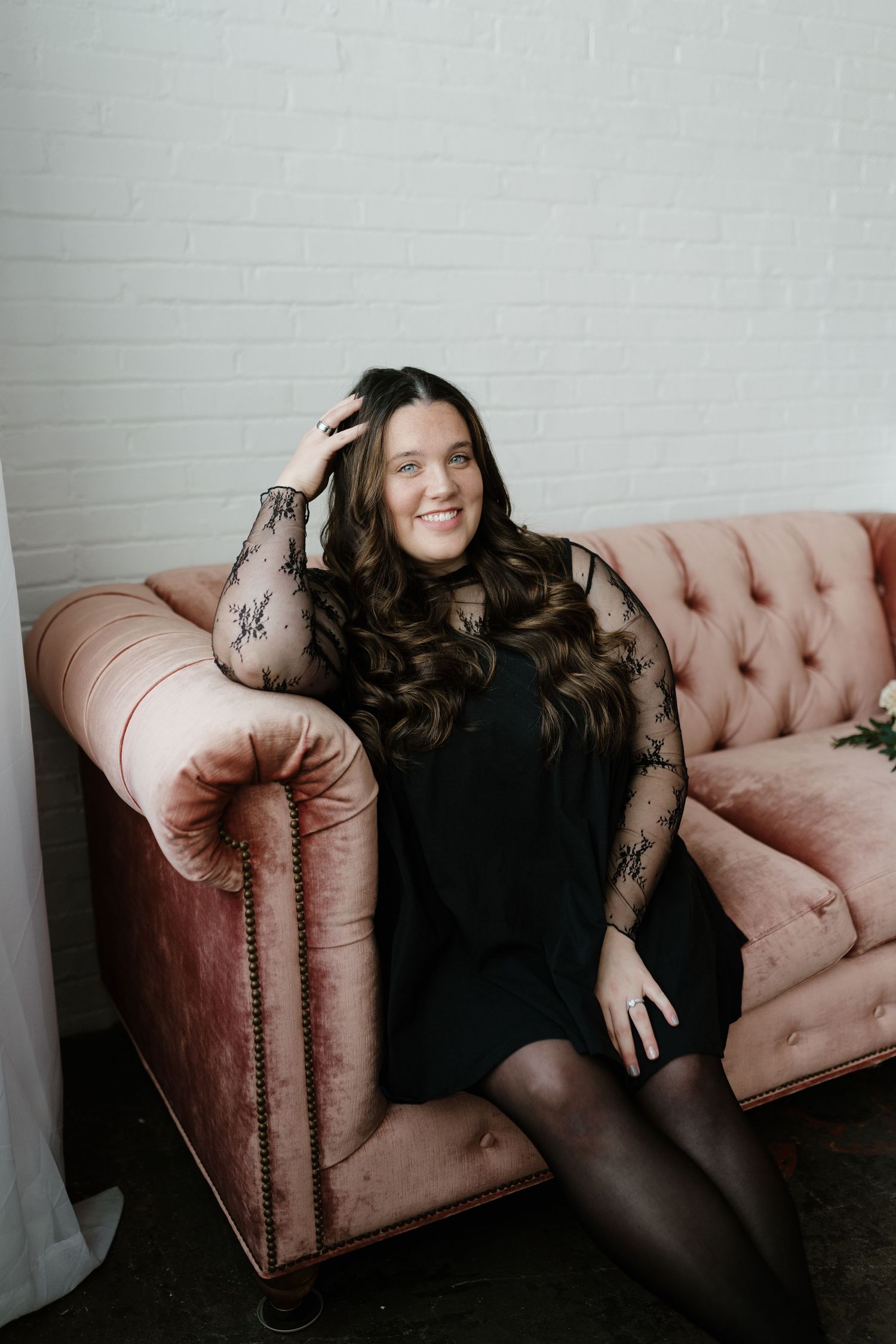 Woman in black lace dress smiling while seated on pink tufted velvet sofa.