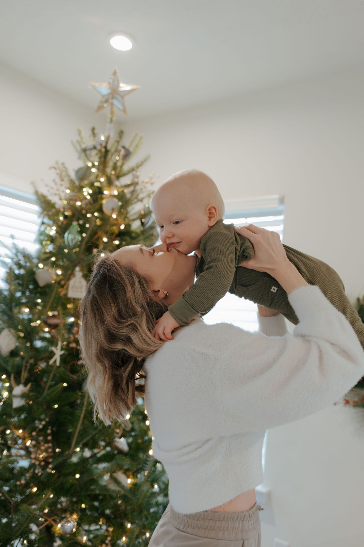 A joyful mother lifts her baby toward her face in front of a decorated Christmas tree.