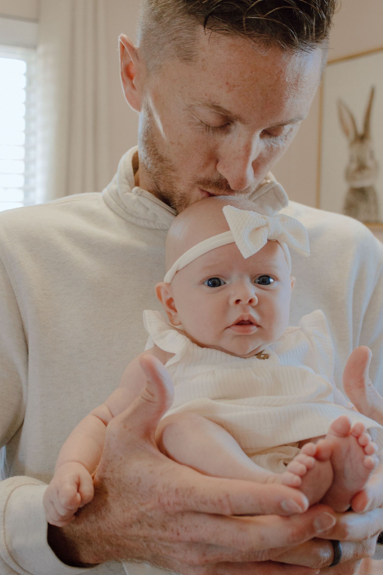 Devoted father kisses the head of his alert newborn daughter dressed in white with a bow headband.