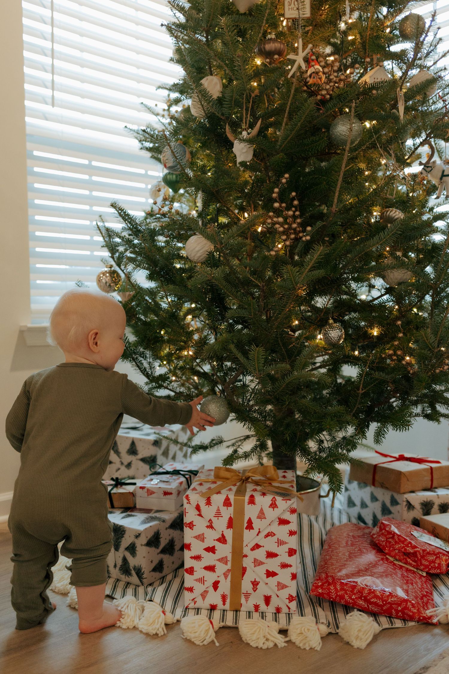 Baby in green outfit stands at base of decorated Christmas tree surrounded by wrapped presents.