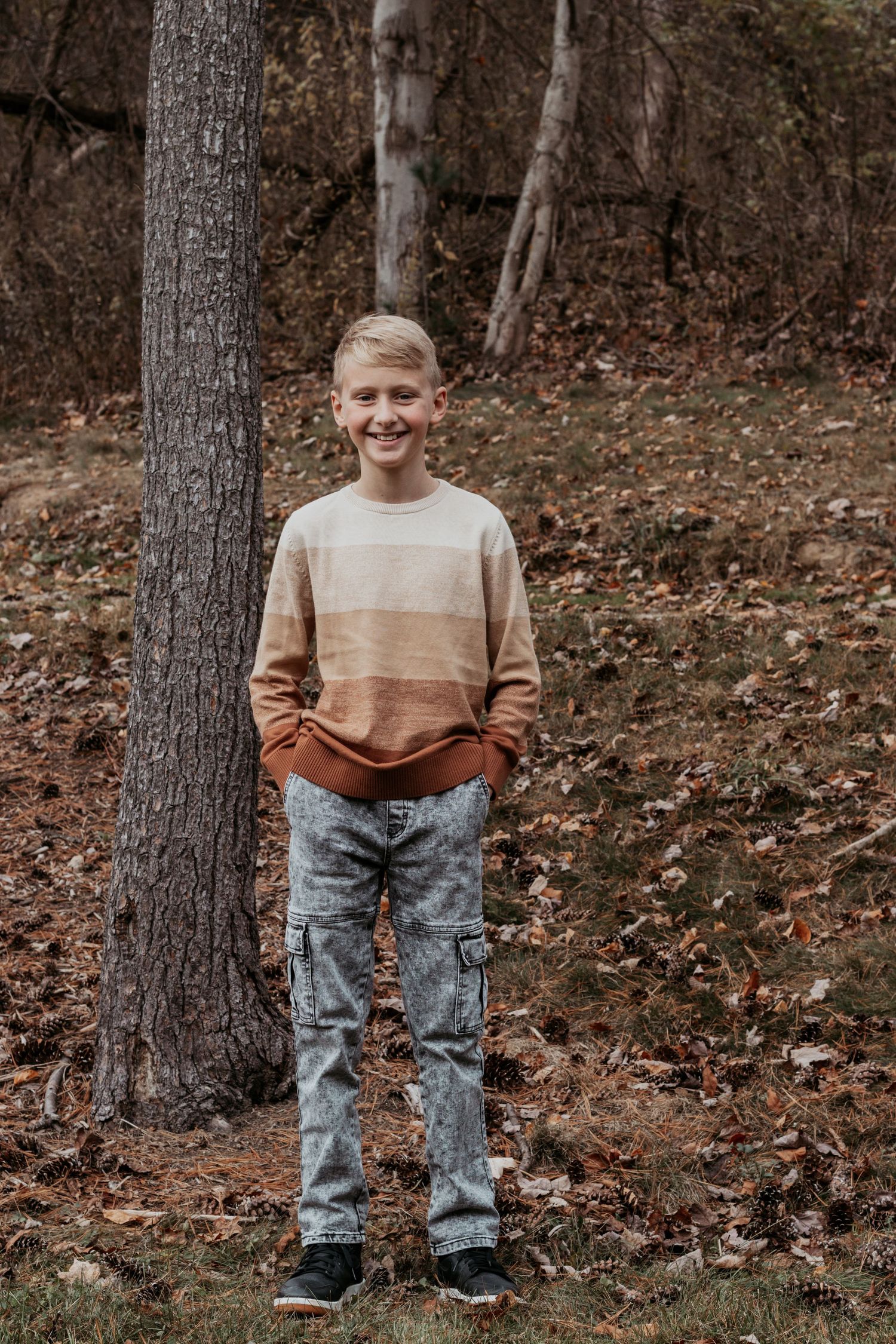 Smiling boy in striped sweater standing by tree trunk in autumn leaves.