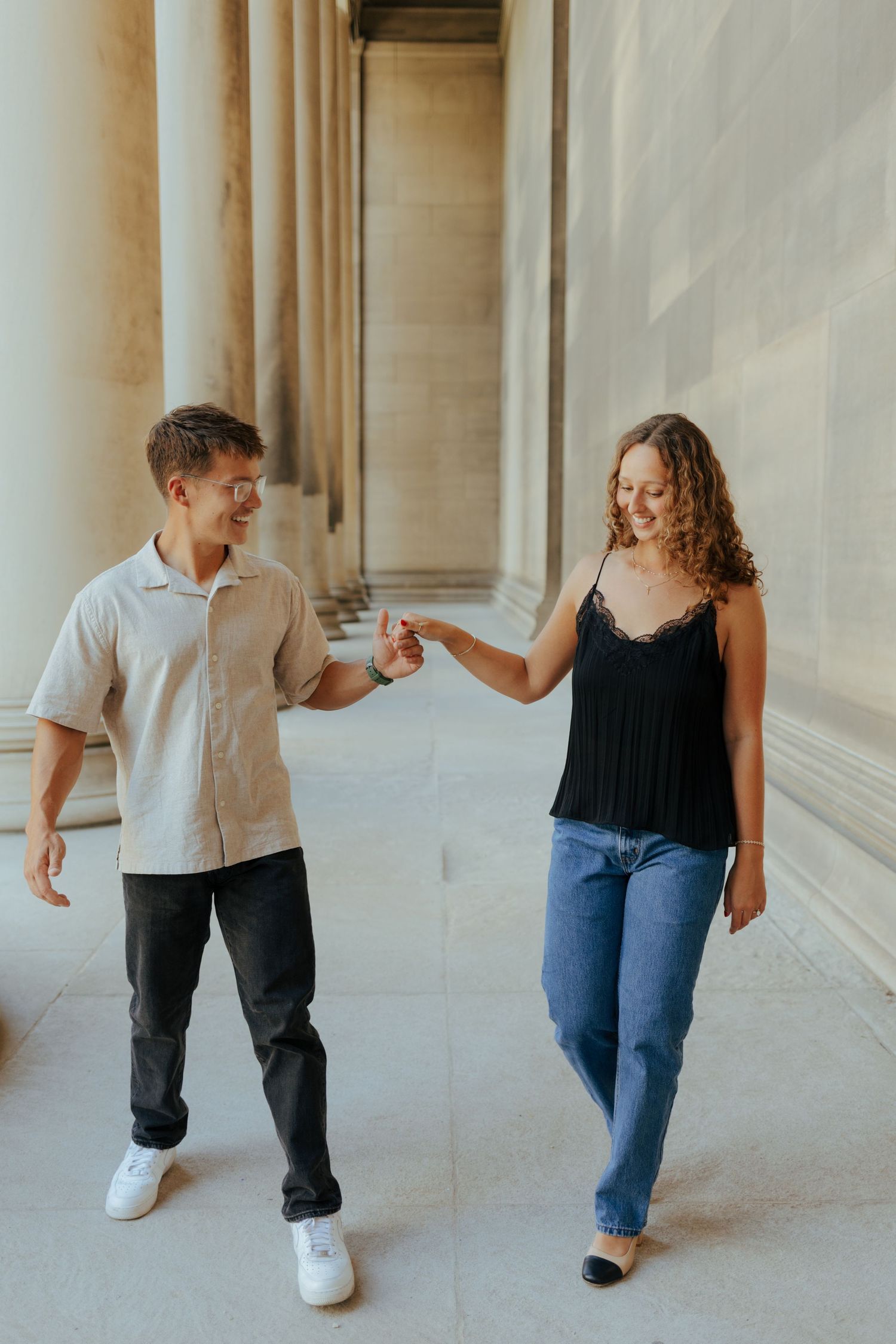 An engaged couple holds hands and smiles while walking between tall stone columns.