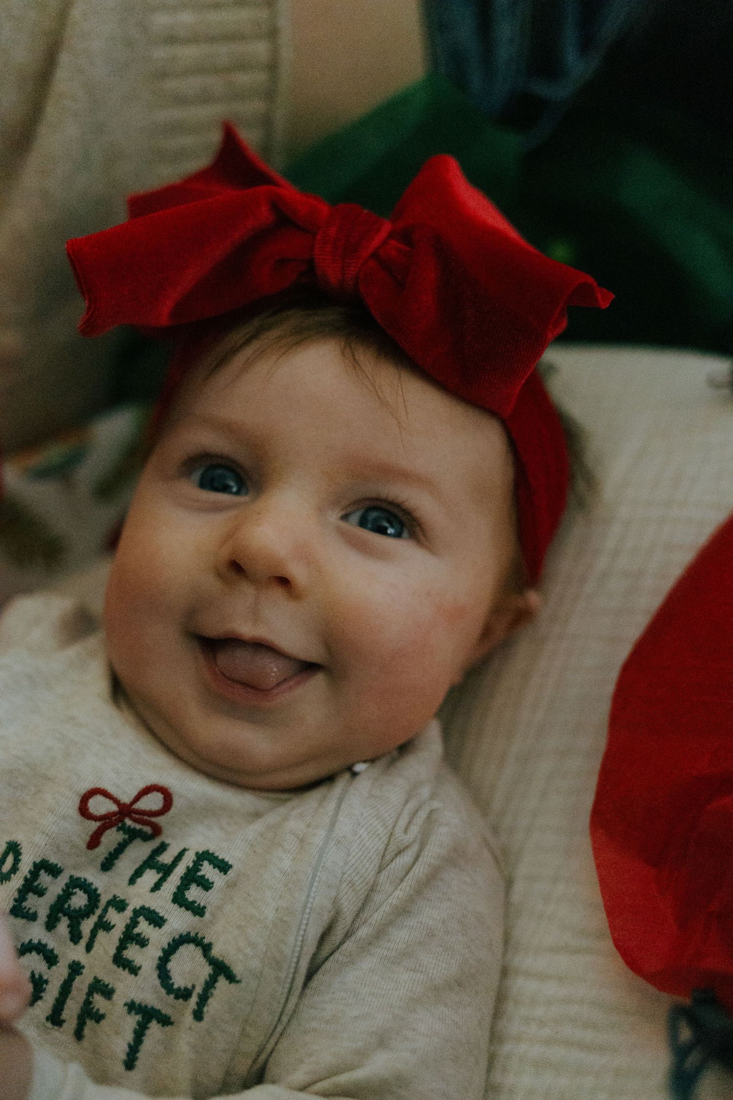 Smiling baby girl with red bow headband wearing The Perfect Gift holiday onesie.