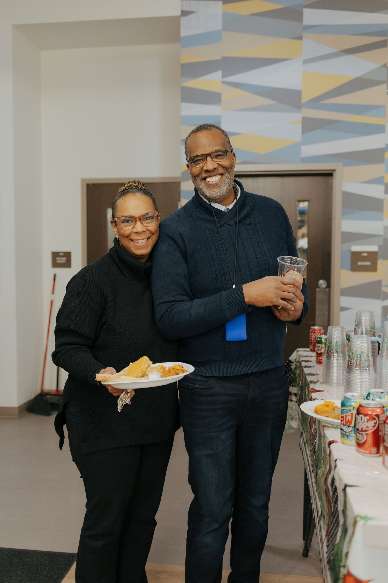 A smiling couple poses together at a holiday party, holding food and drinks near a festive refreshment table.