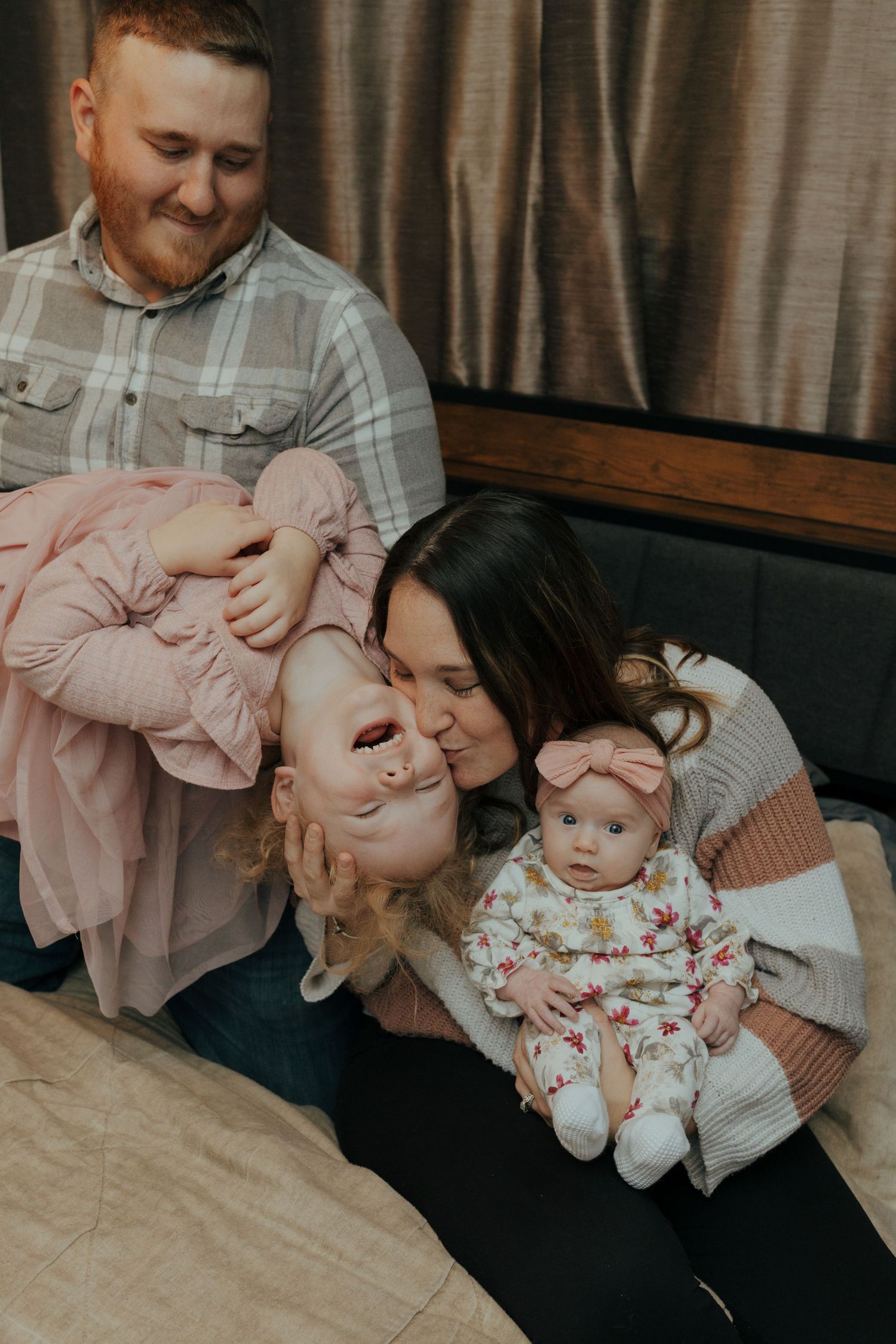 Joyful family with father, mother kissing toddler, and newborn baby sitting together on bed.
