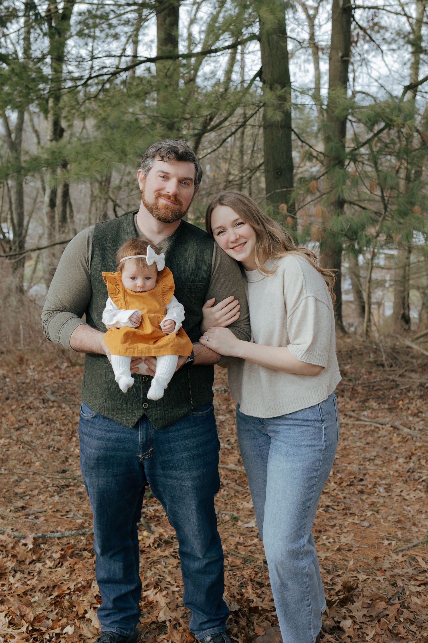 Young family with bearded father holding baby girl in orange dress in autumn woods.