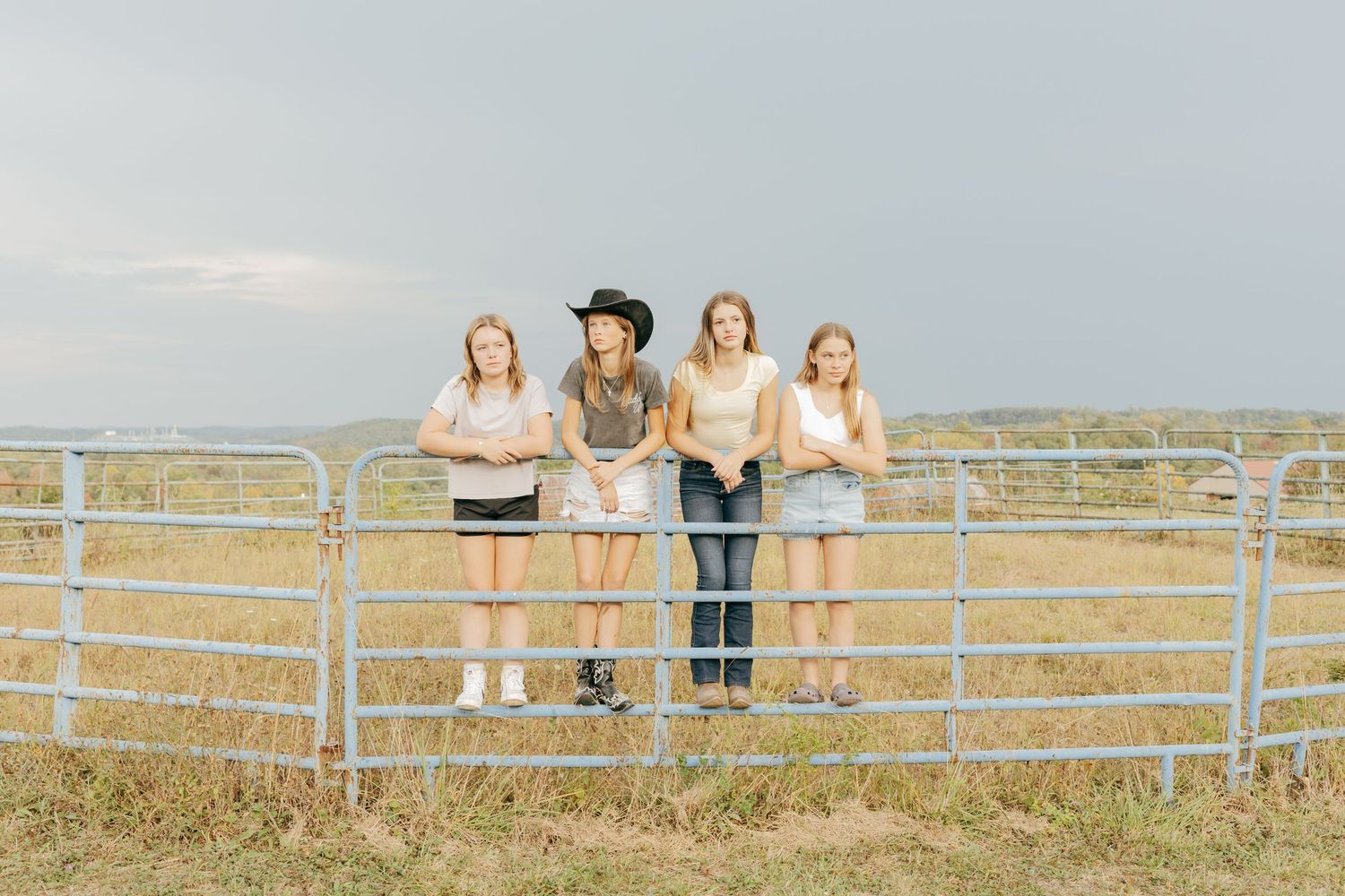 Four girls pose at a farm gate in a rural field under a cloudy sky.