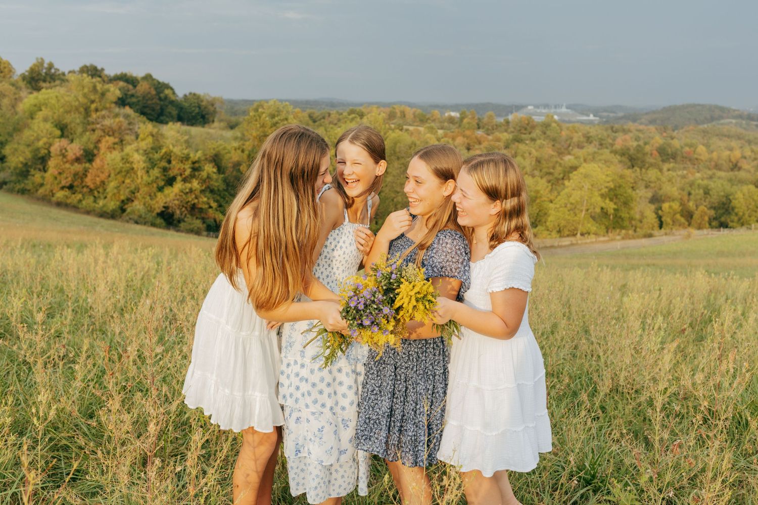 Four girls in summer dresses sharing a bouquet of yellow wildflowers in a meadow.