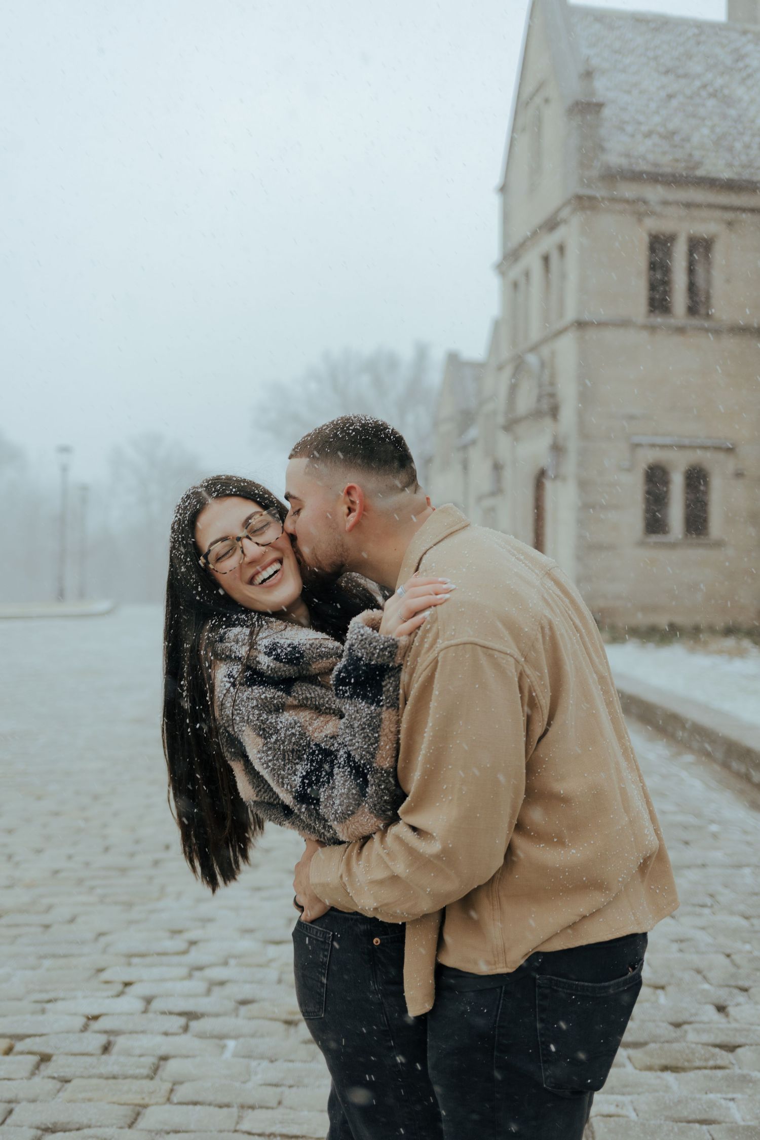 Couple laughing and embracing in falling snow outside a historic stone building.