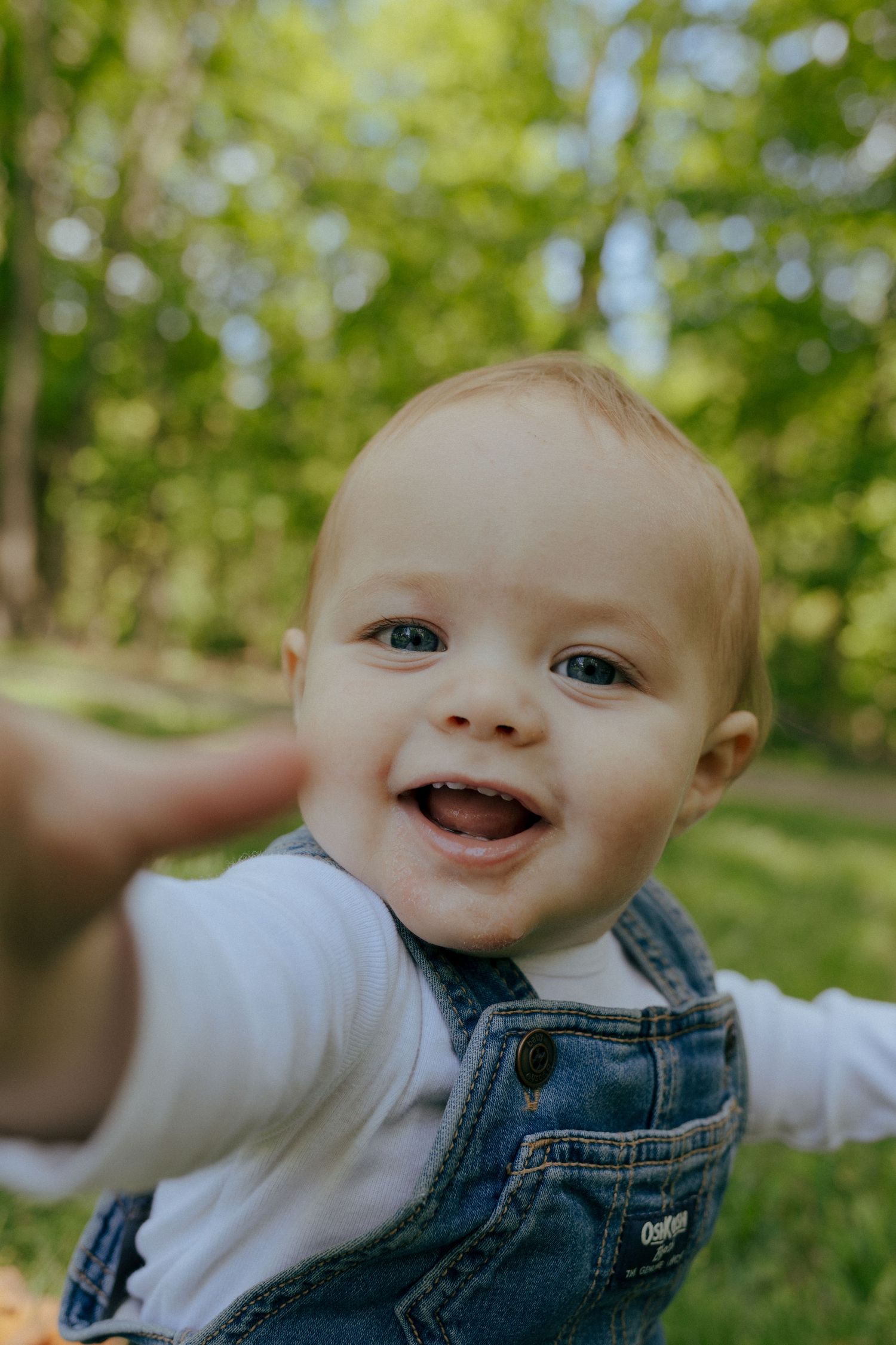 Smiling baby in denim OshKosh overalls reaching toward camera in a lush green outdoor setting.