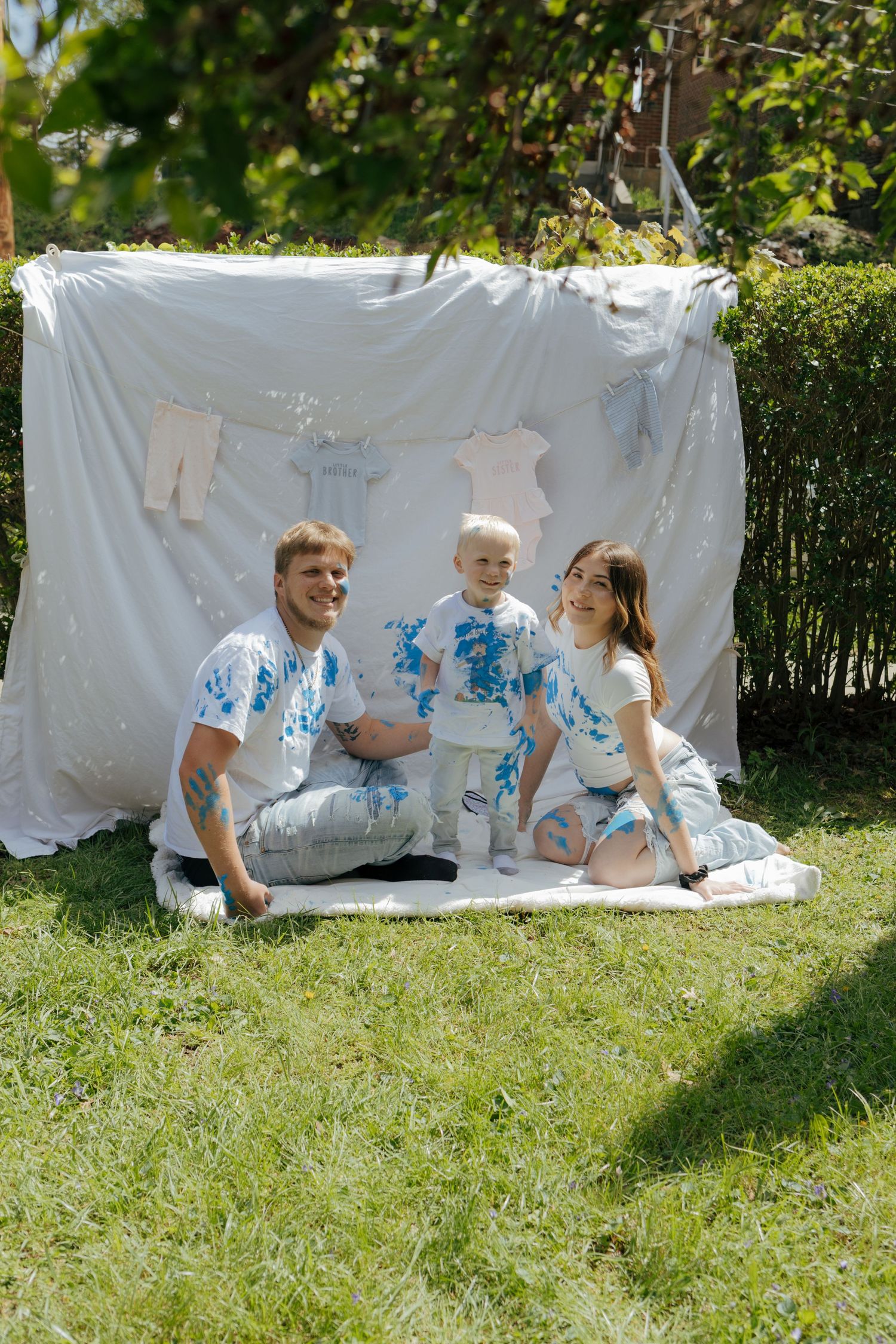Family of three sitting on grass with blue paint on white outfits in front of a white backdrop outdoors.