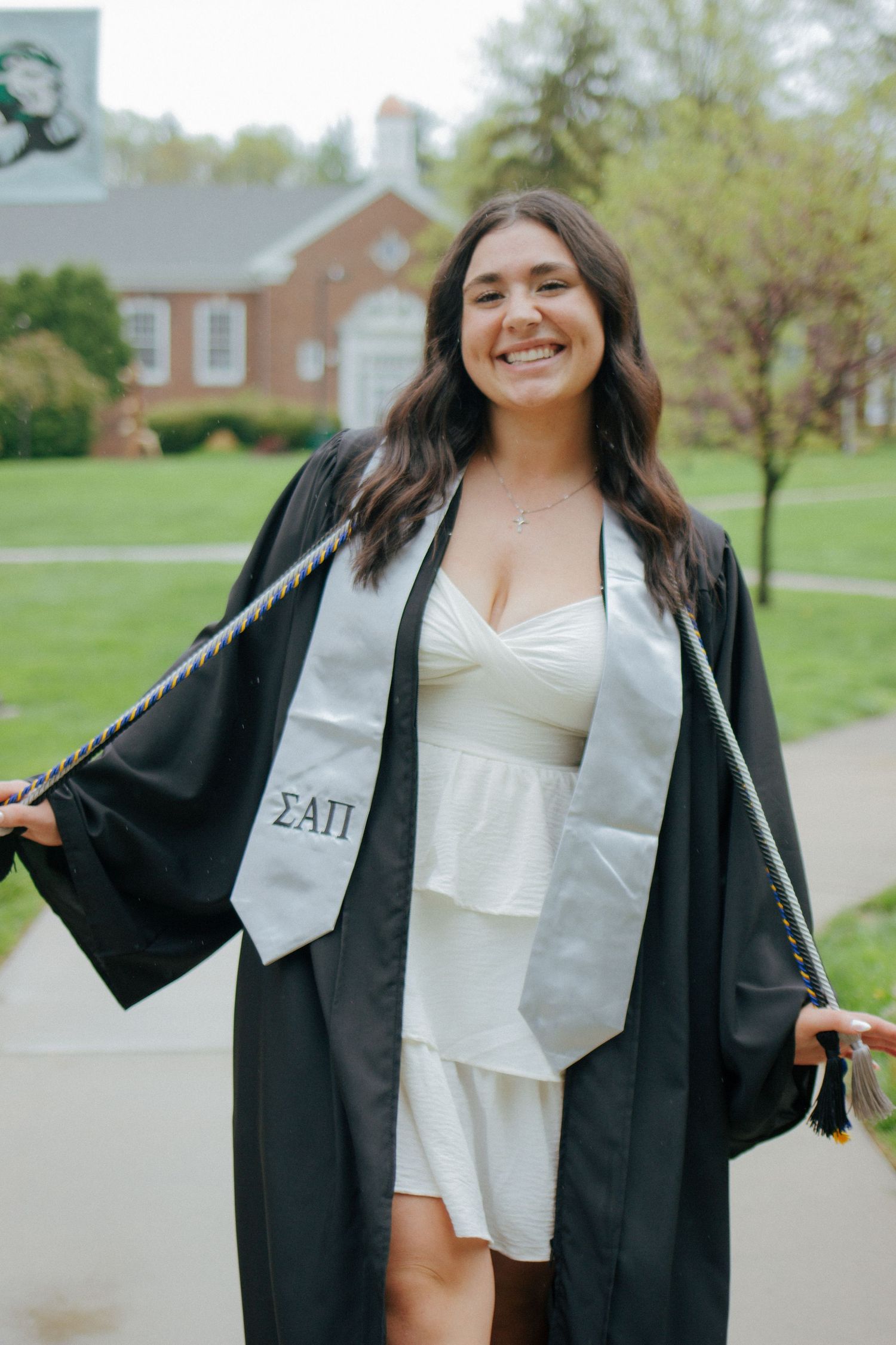 Smiling college graduate in black gown and Sigma Alpha Pi stole posing on a green campus lawn.