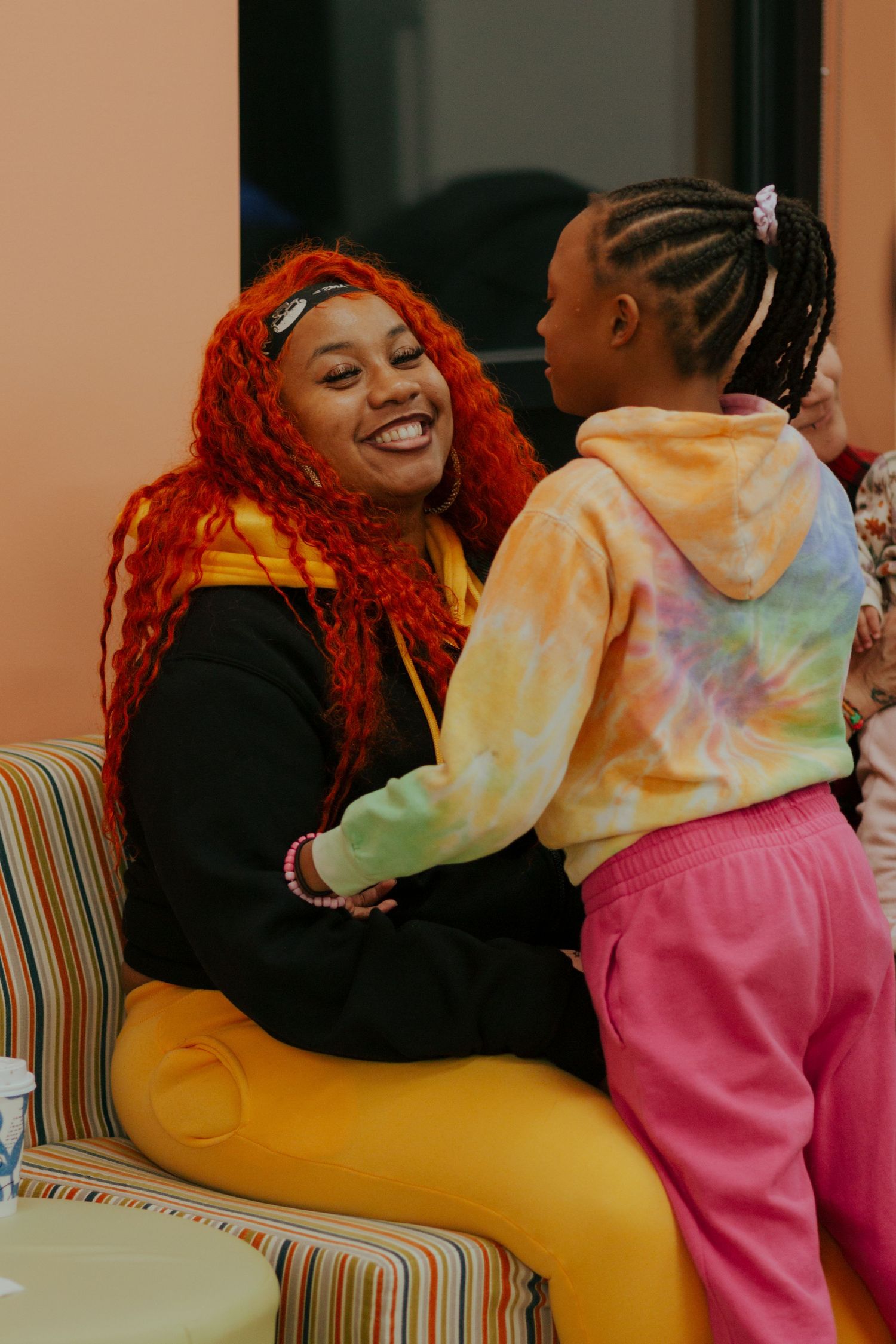 A smiling woman with vibrant red locs shares a joyful moment with a young girl in a colorful tie-dye hoodie.