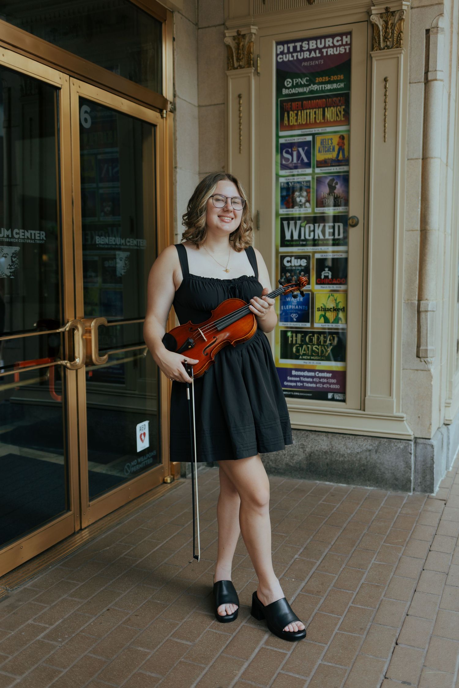Smiling young woman in black dress holding violin outside Pittsburgh Cultural Trust theater.