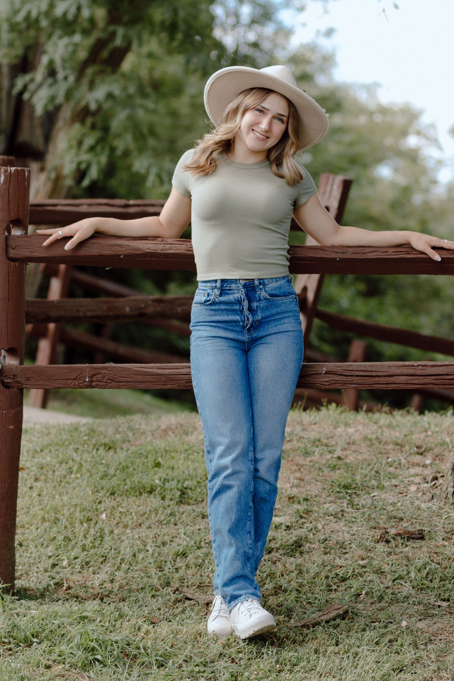 Blonde teen in sage top and wide-brim hat leaning on rustic wooden fence outdoors.