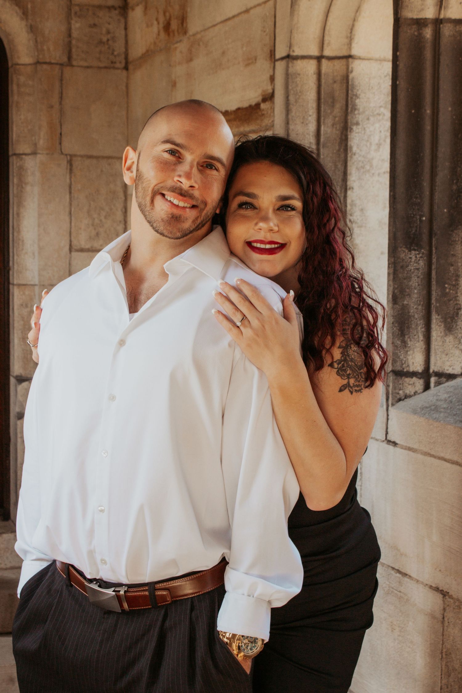 Smiling couple embracing against stone wall, man in white shirt, woman in black dress.