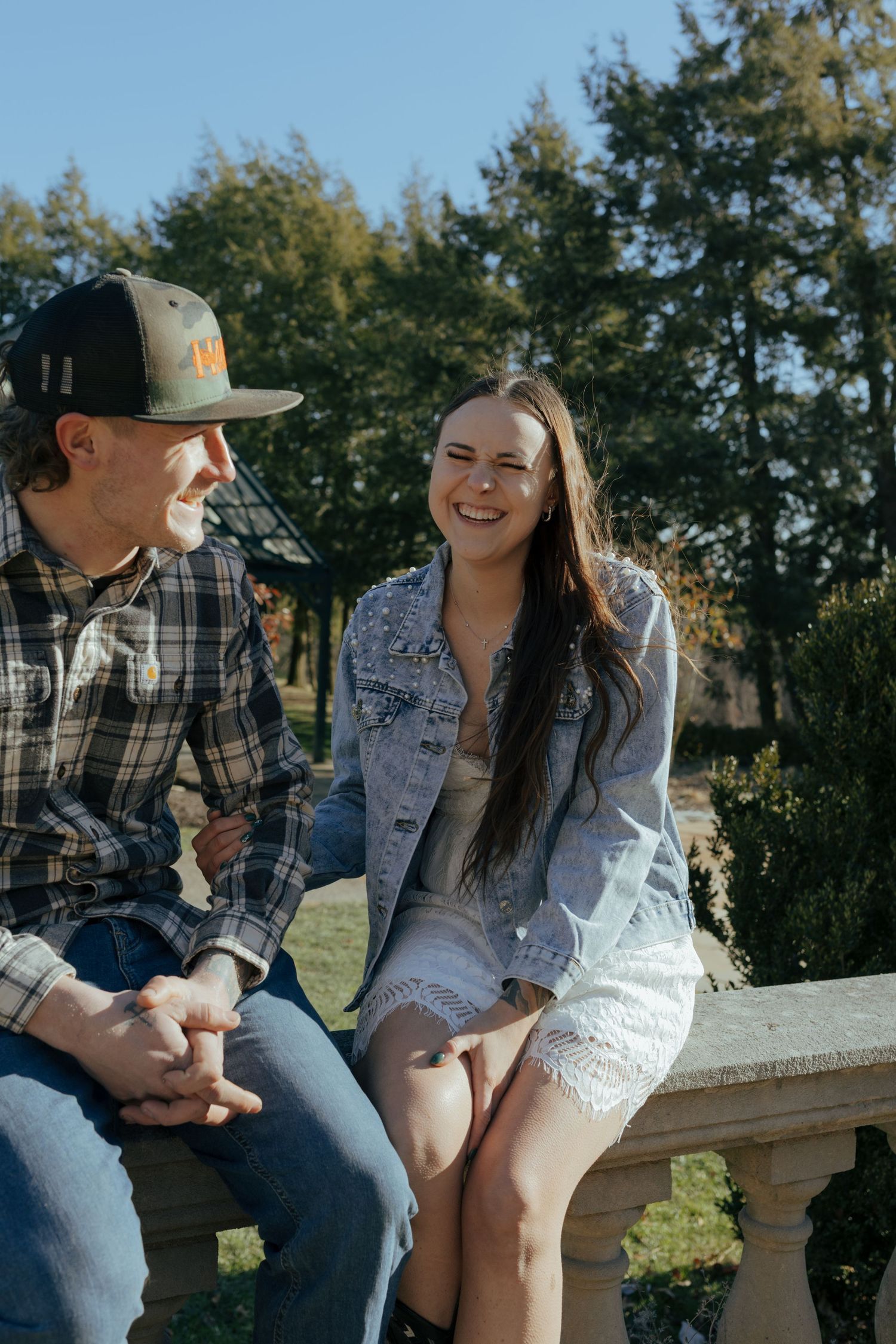 A couple sits on a stone railing outdoors, smiling and laughing together on a sunny day.