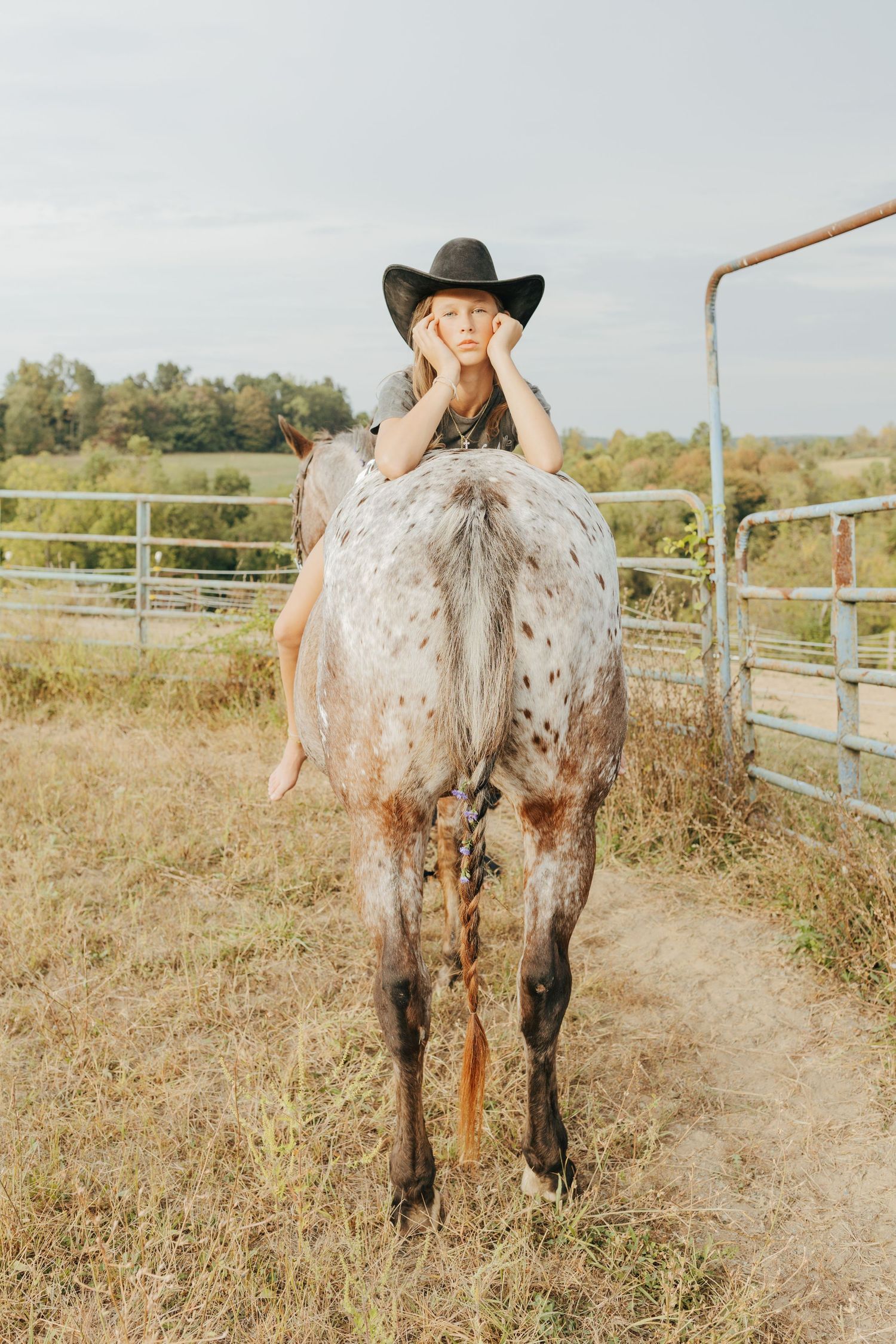 Girl in black cowboy hat leans over a spotted horse in a farm paddock.