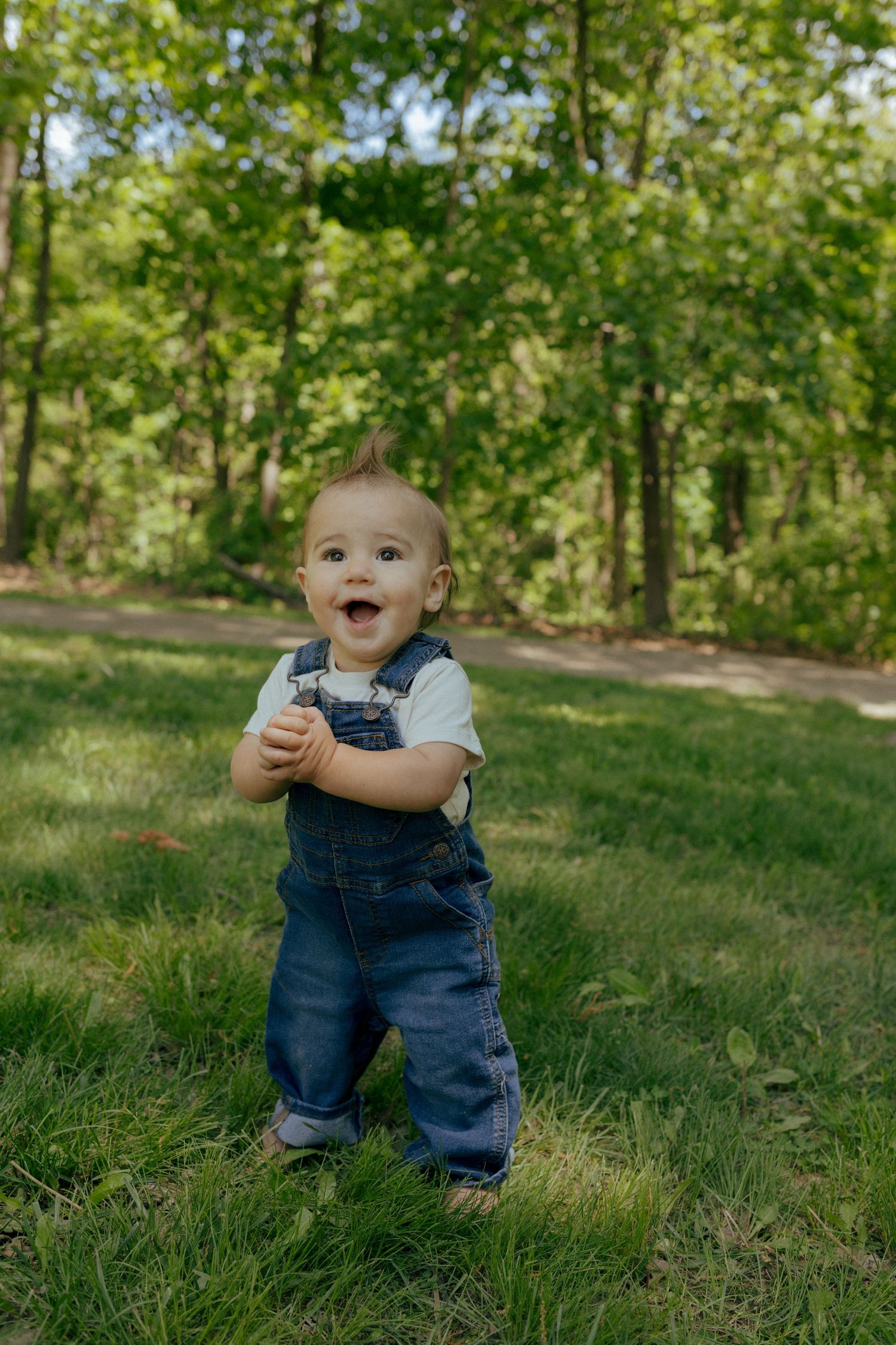 Happy baby in denim overalls standing on green grass in a wooded park, smiling joyfully.