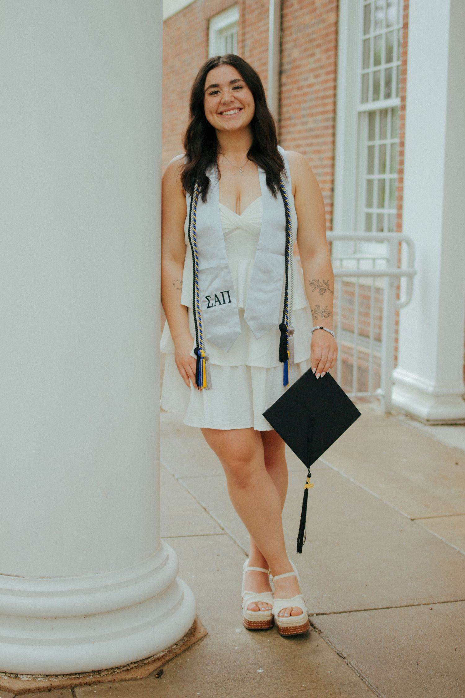 Smiling graduate in white dress holds cap with sorority sash outdoors near brick building columns.