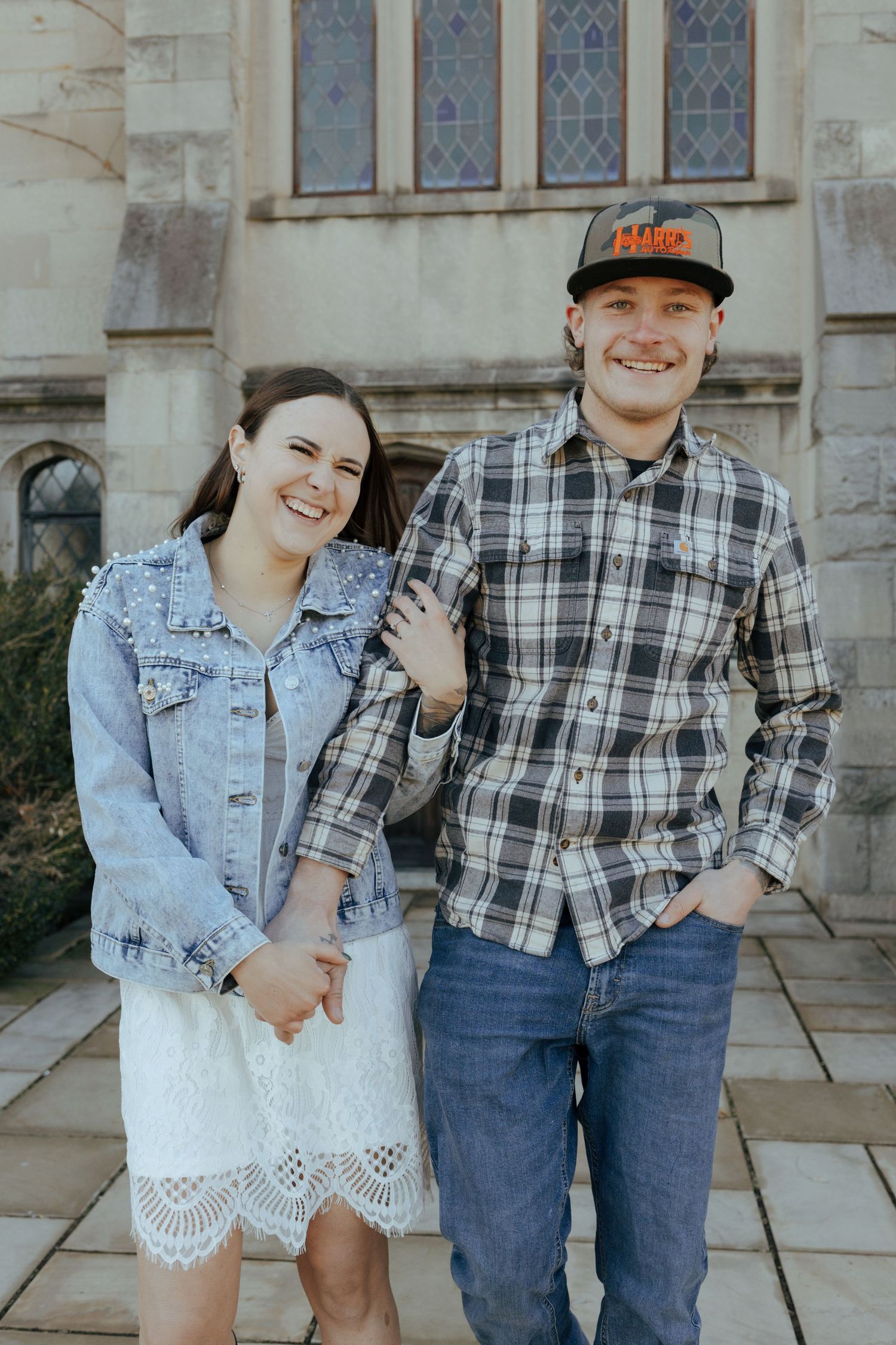 Smiling engaged couple holds hands posing together outside gothic stone church building.