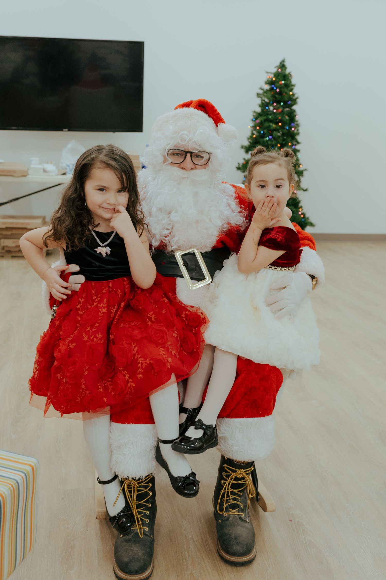 Two young girls in elegant red dresses pose with Santa Claus in front of a decorated Christmas tree.
