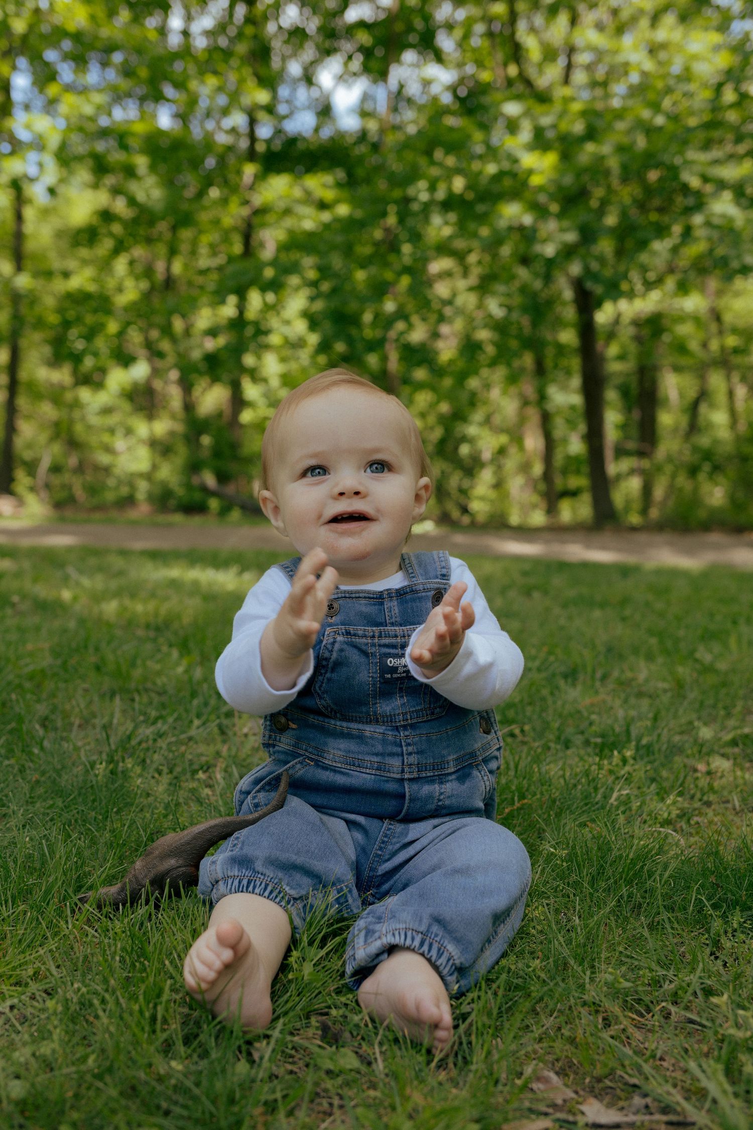 Adorable baby in denim overalls sitting on green grass in a sunny park, clapping hands.