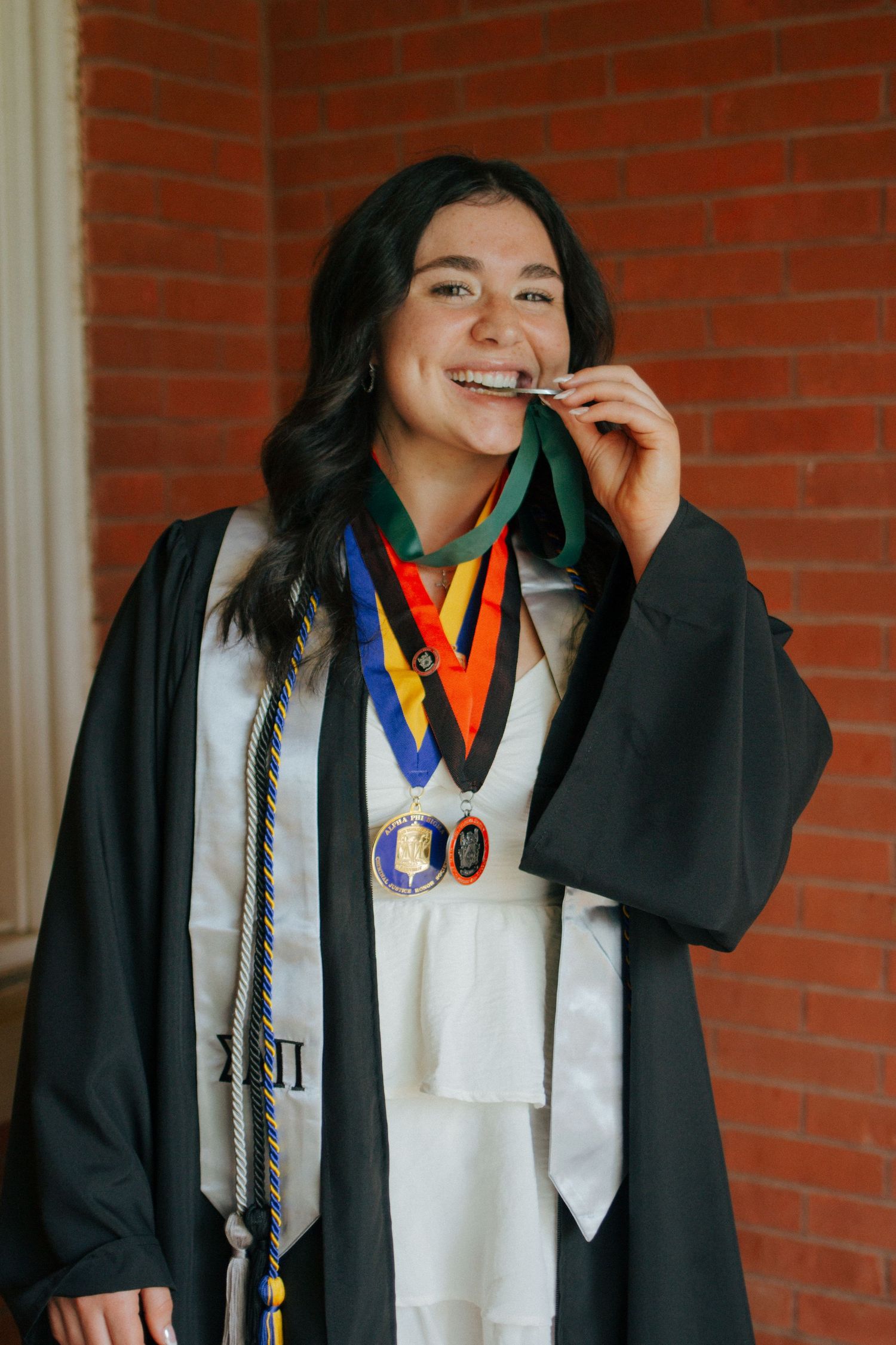 Proud graduate in cap and gown biting her diploma while wearing multiple honor medals and cords.