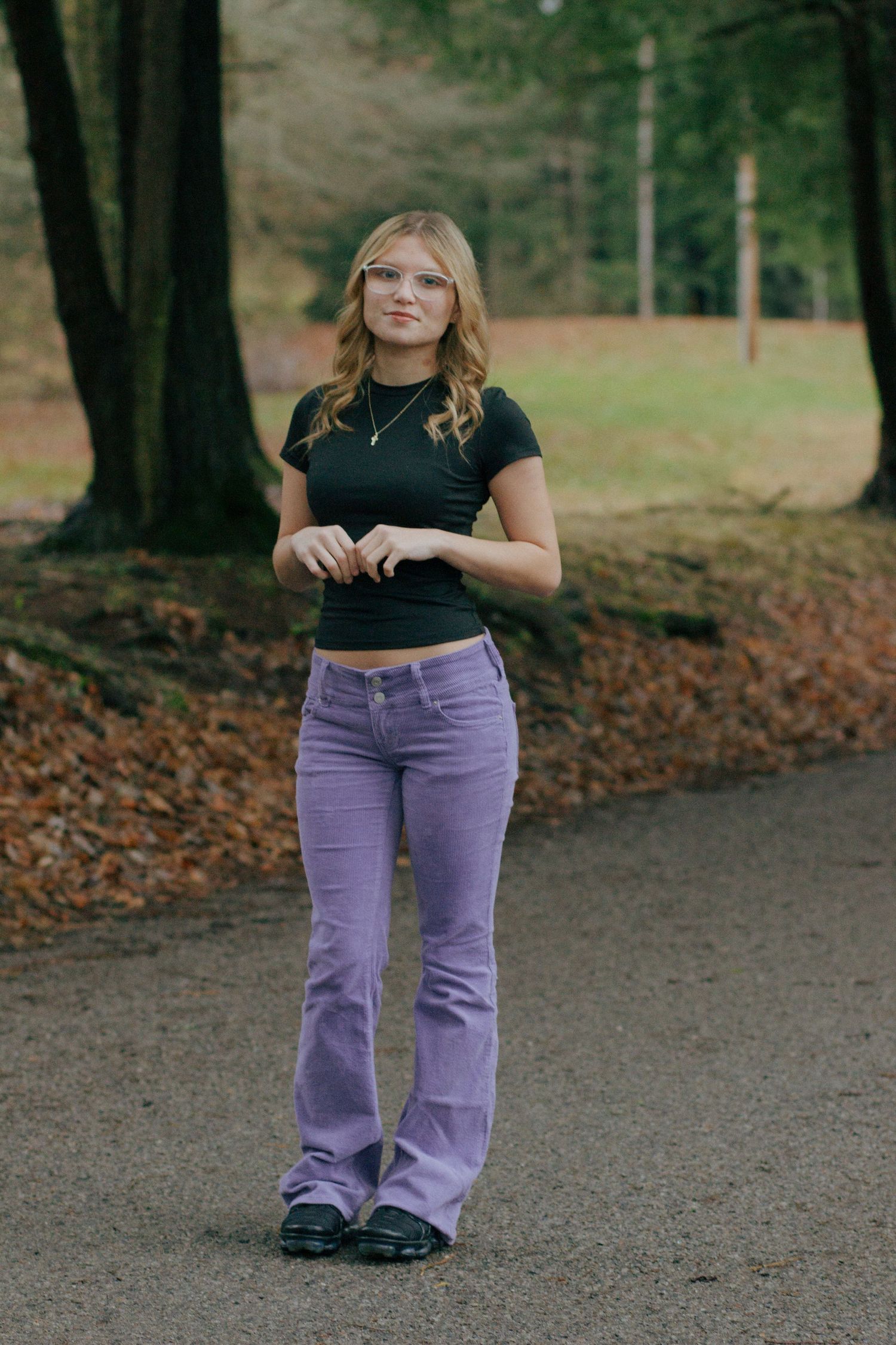 Blonde teen girl in black crop top and purple flare jeans posing on a park path.
