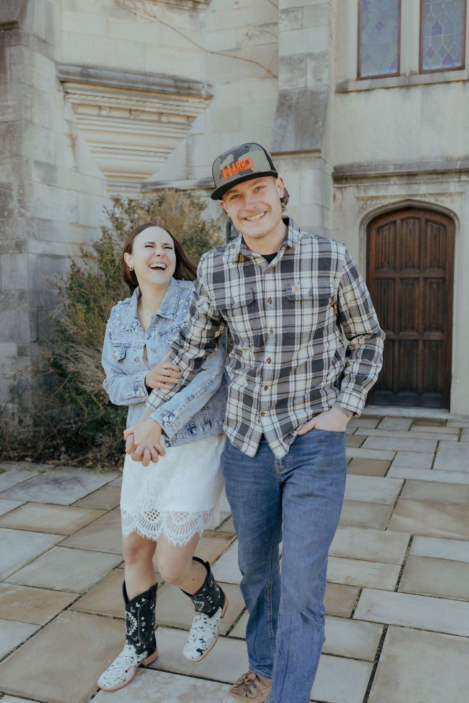 Laughing couple walks hand in hand outside a stone building on a sunny day.