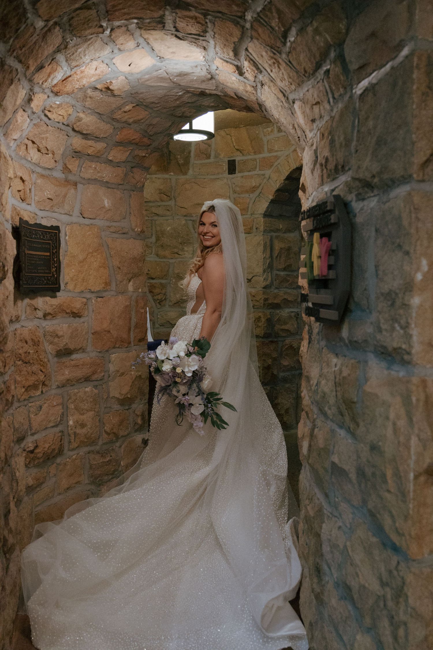 Elegant bride in a pearl wedding gown and veil holding a bouquet in a stone archway corridor.