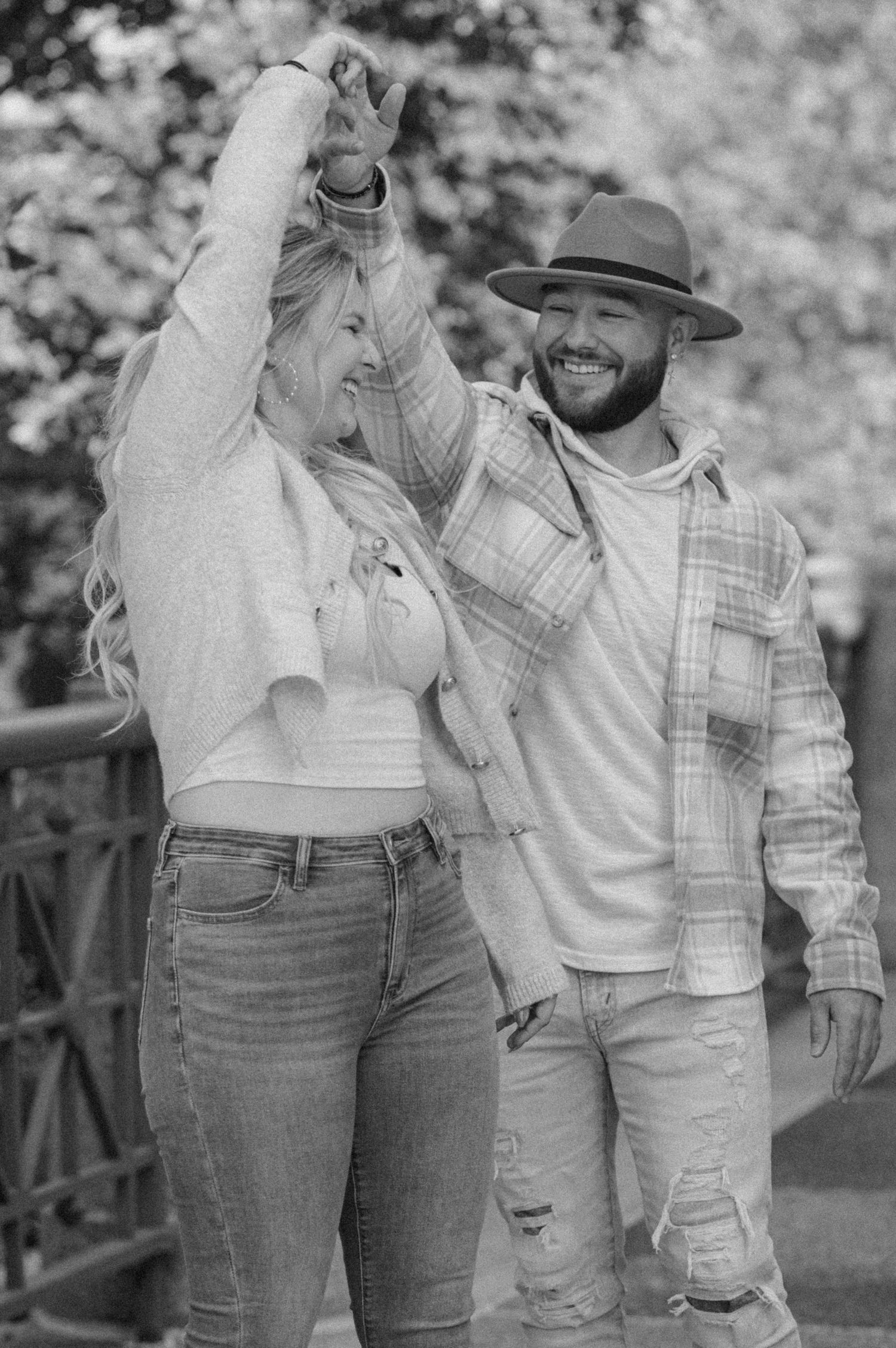 Happy couple dancing outdoors in black and white photo, man wearing a hat and woman with long blonde hair.