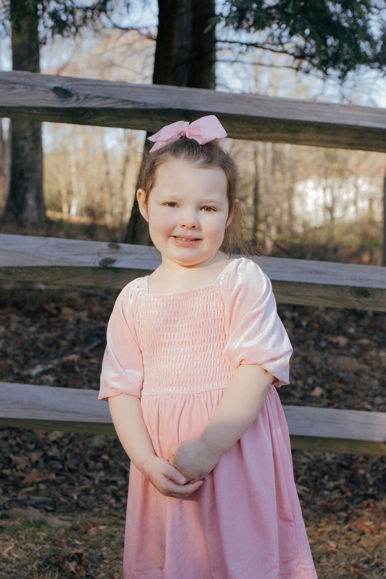 Cute young girl in pink smocked dress with matching bow smiles in front of a wooden fence.