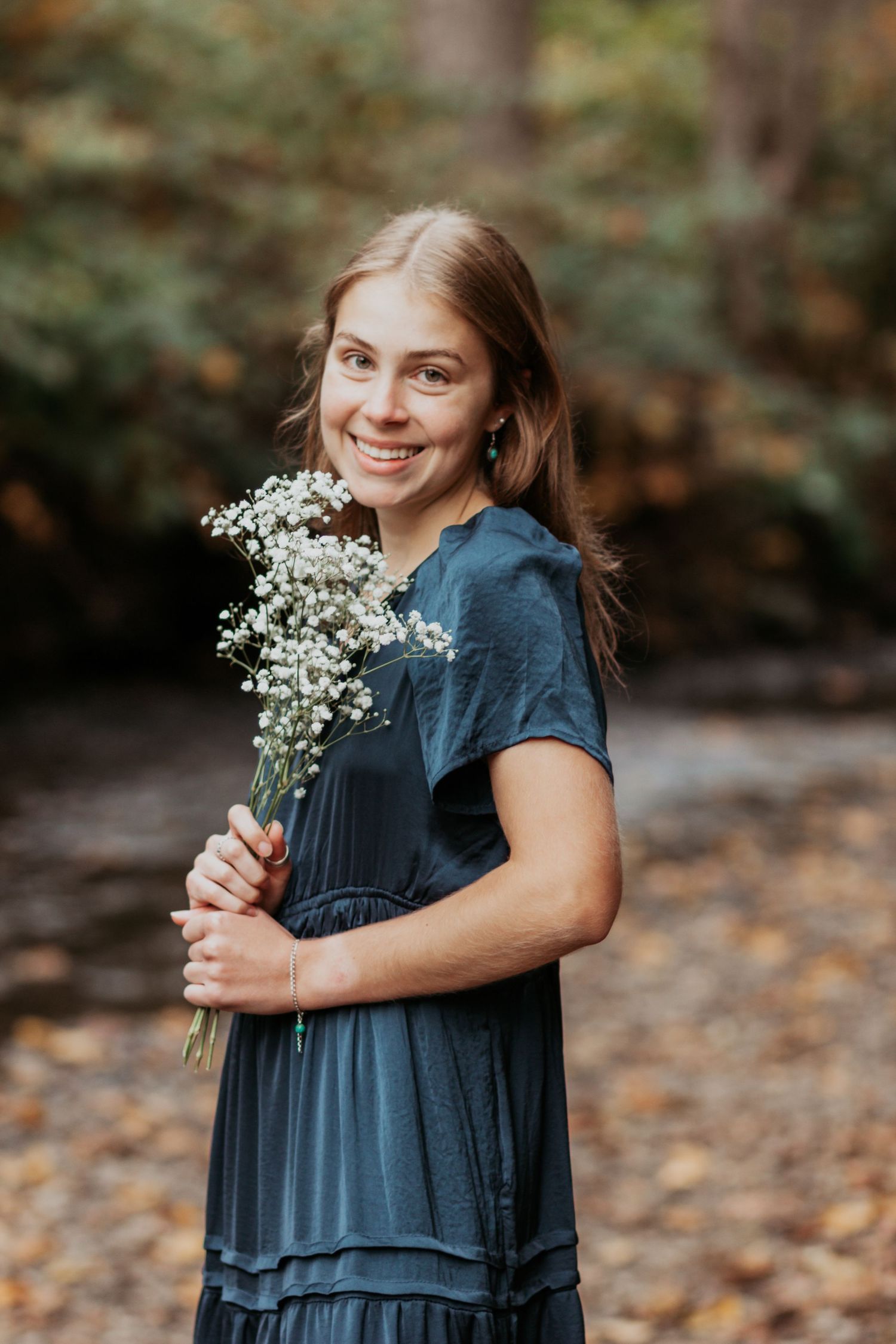 Smiling young woman in dark blue dress holds white wildflower bouquet in autumn forest setting.