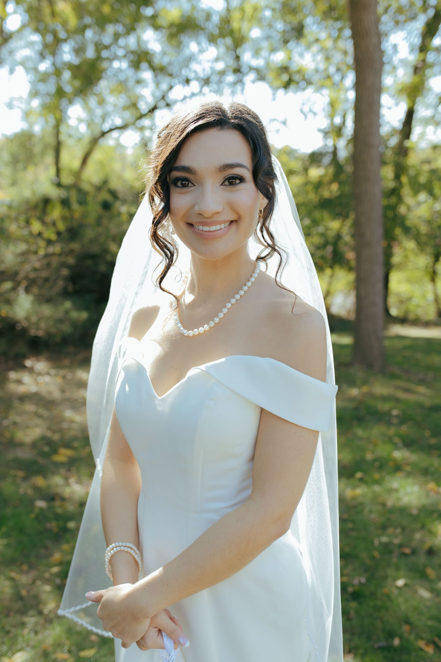 Smiling bride in off-shoulder white gown with pearl necklace posing outdoors in park.