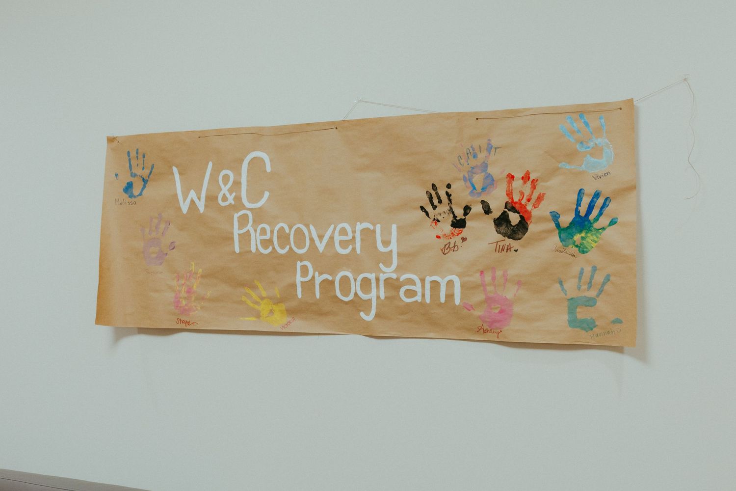 A handmade banner decorated with colorful handprints reads W&C Recovery Program on a white wall.